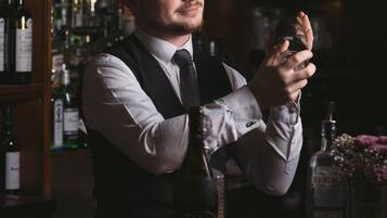 A bartender in a vest and tie holding a glass. There are bottles and a bar counter in the background.