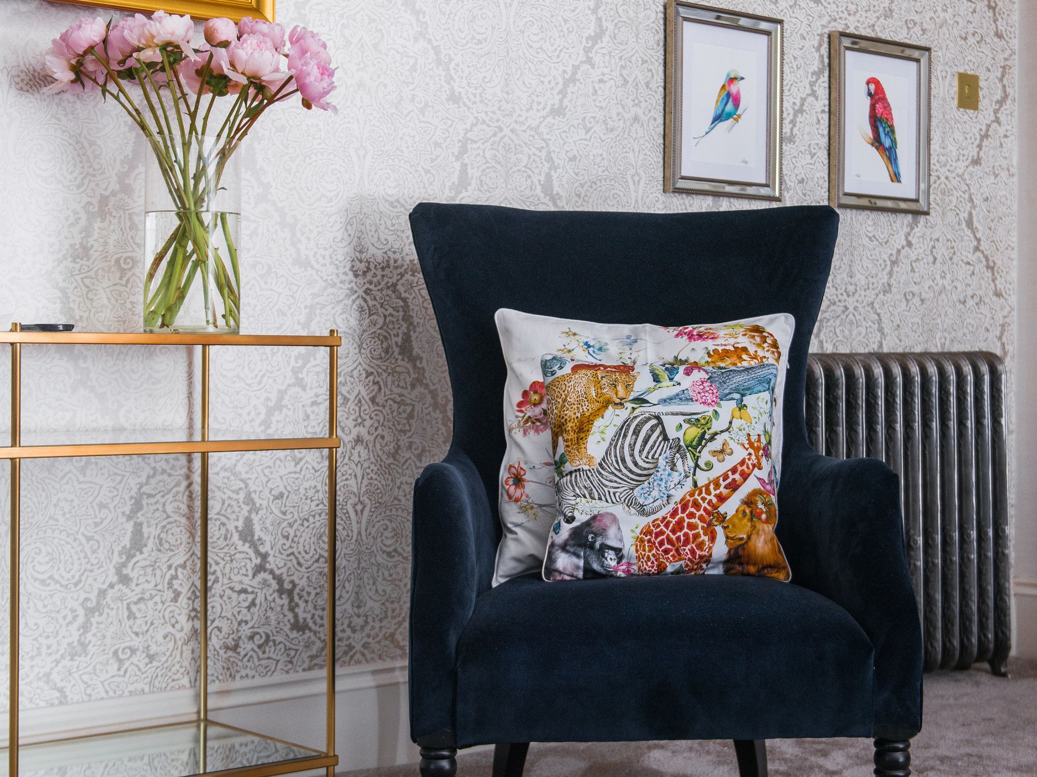 A dark blue armchair with a colorful animal print pillow, a gold-framed picture frame, and a vase with pink flowers on a gold stand.