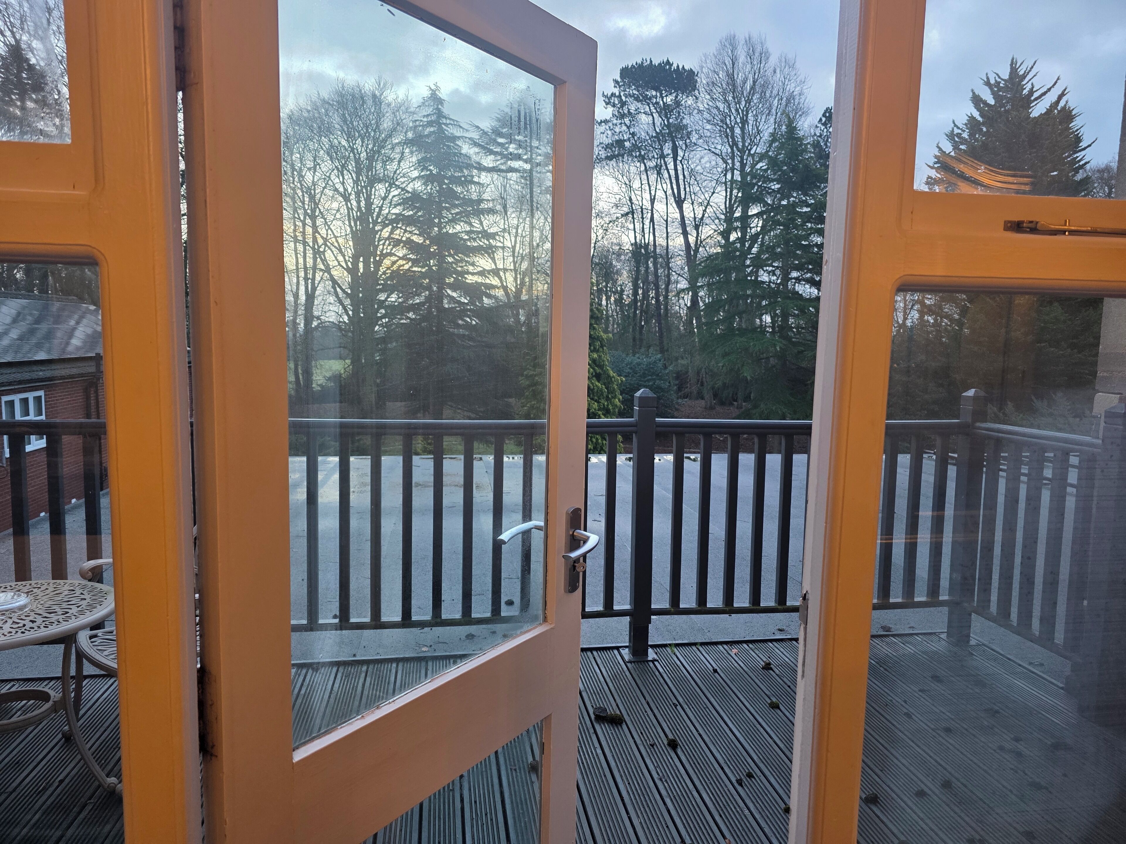 A view from a balcony with a wooden deck, a metal railing, and a glass door leading to a patio area with a table and chairs.