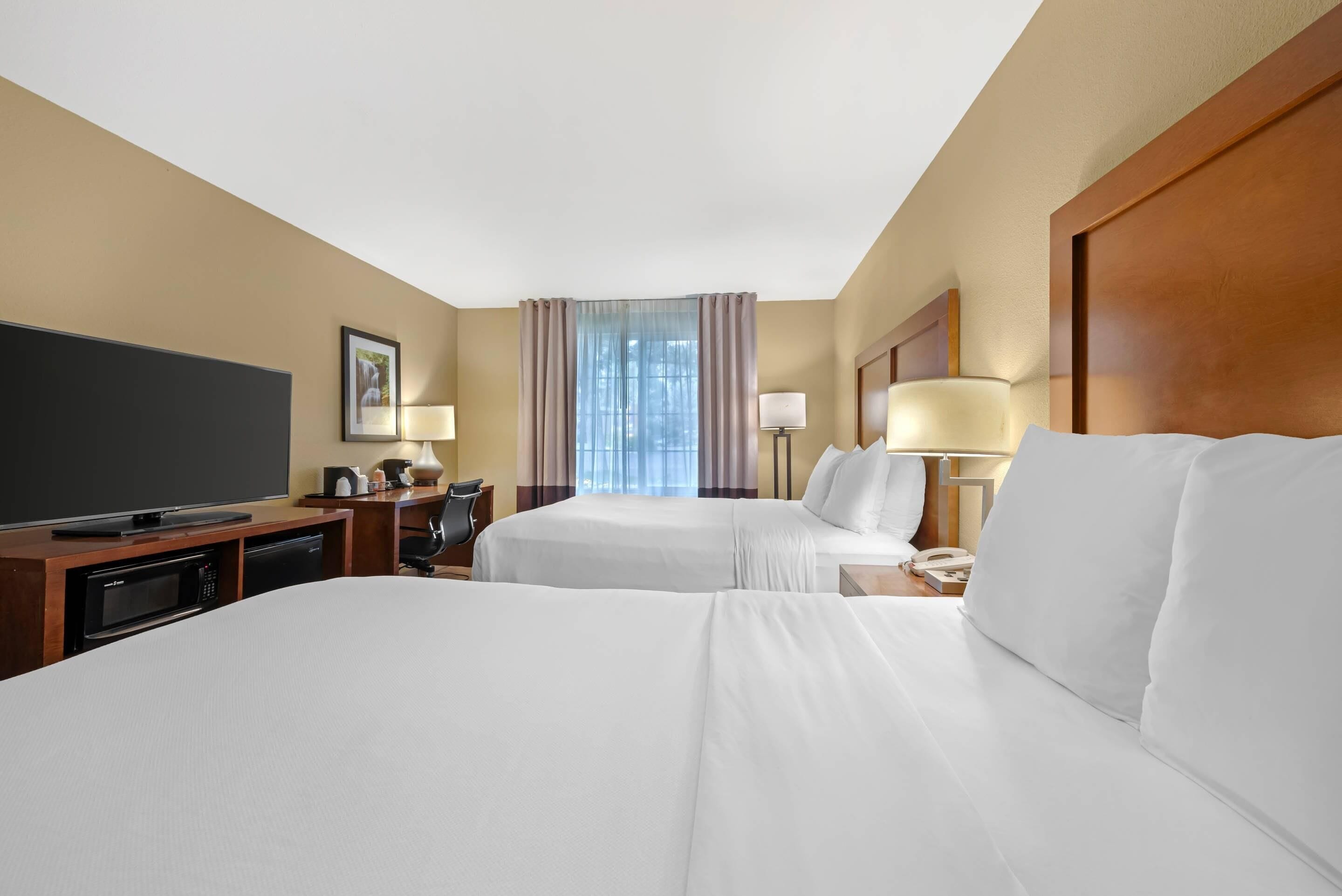 A hotel room with two beds, a television, a desk with a lamp, and a window with curtains.