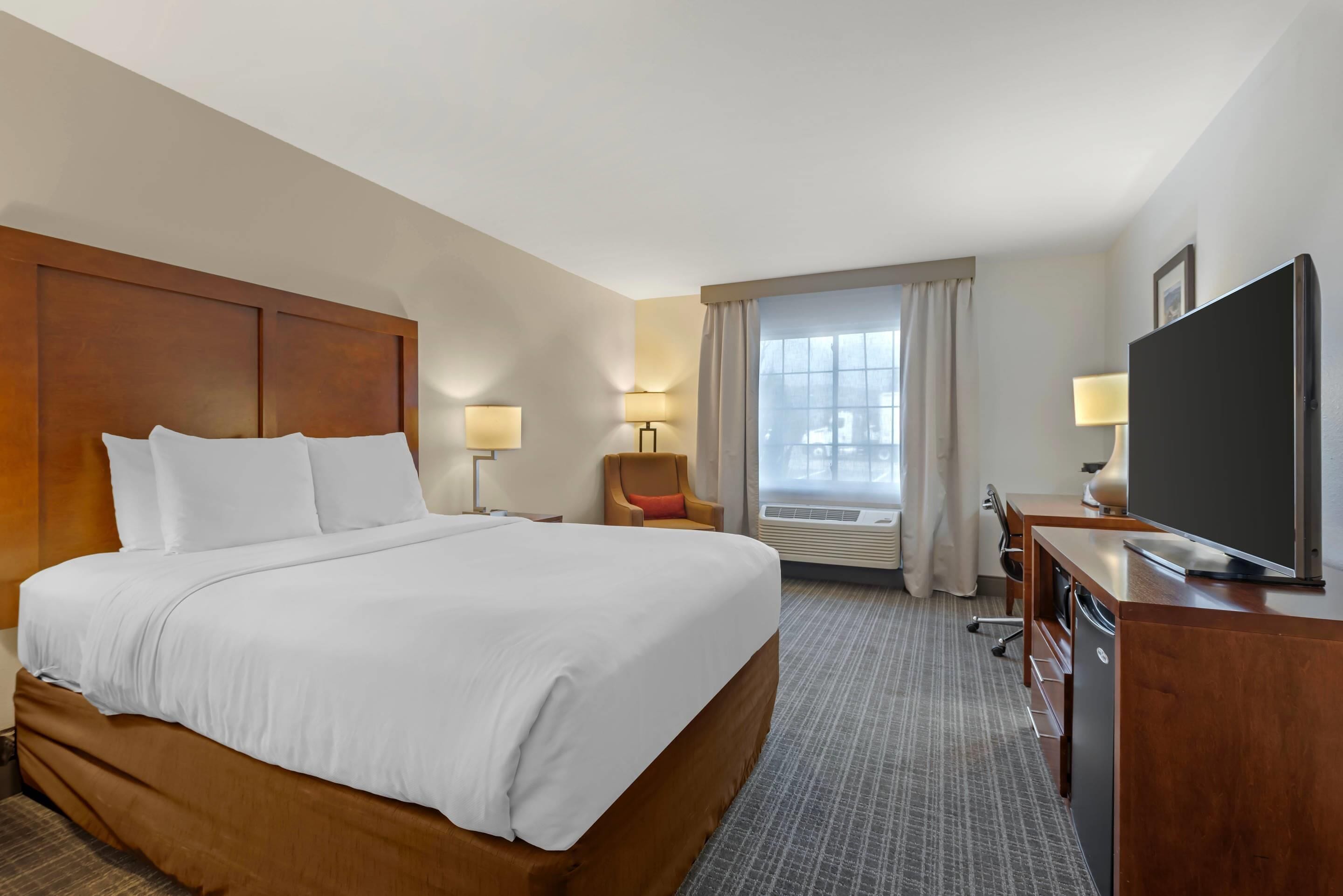 Ein Hotelzimmer mit einem großen Bett, einem Schreibtisch mit Fernseher, einem Sessel und einem fenster mit Vorhängen.