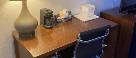 Habitación de hotel con un escritorio de madera, una silla de oficina negra, una lámpara, un cuadro enmarcado de flores, un televisor y un pequeño cesto de basura.