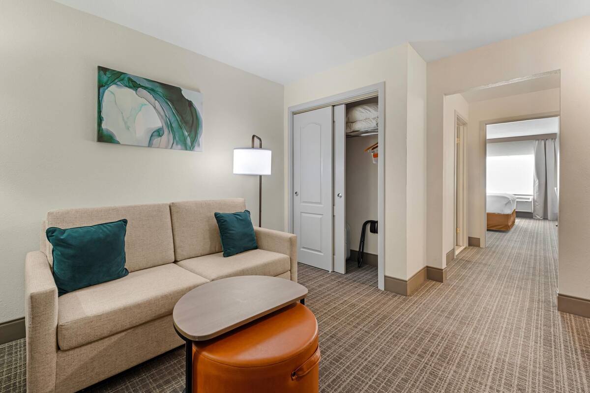 A hotel room with a beige sofa, teal pillows, a small table, a lamp, and a closet with a hanging rack.