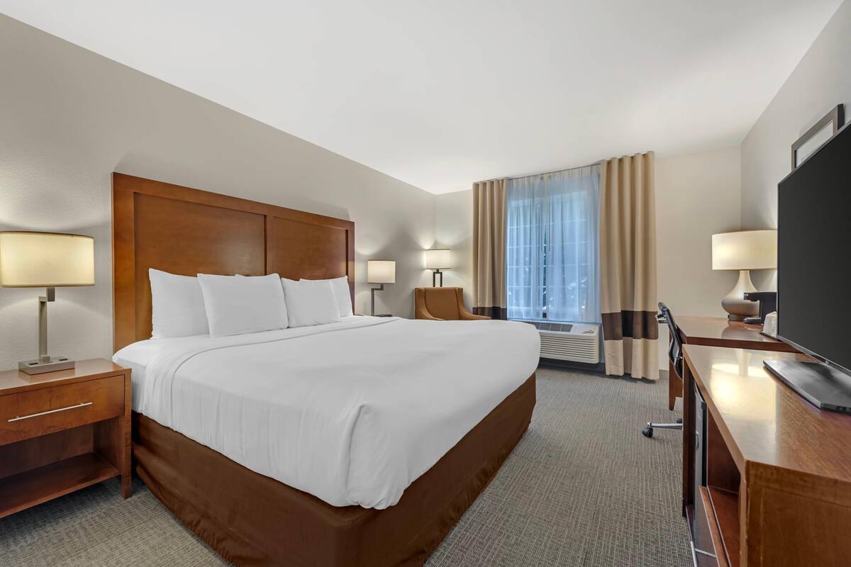 A hotel room with a large bed, two bedside tables with lamps, a desk with a chair, a flat-screen TV, and a window with curtains.
