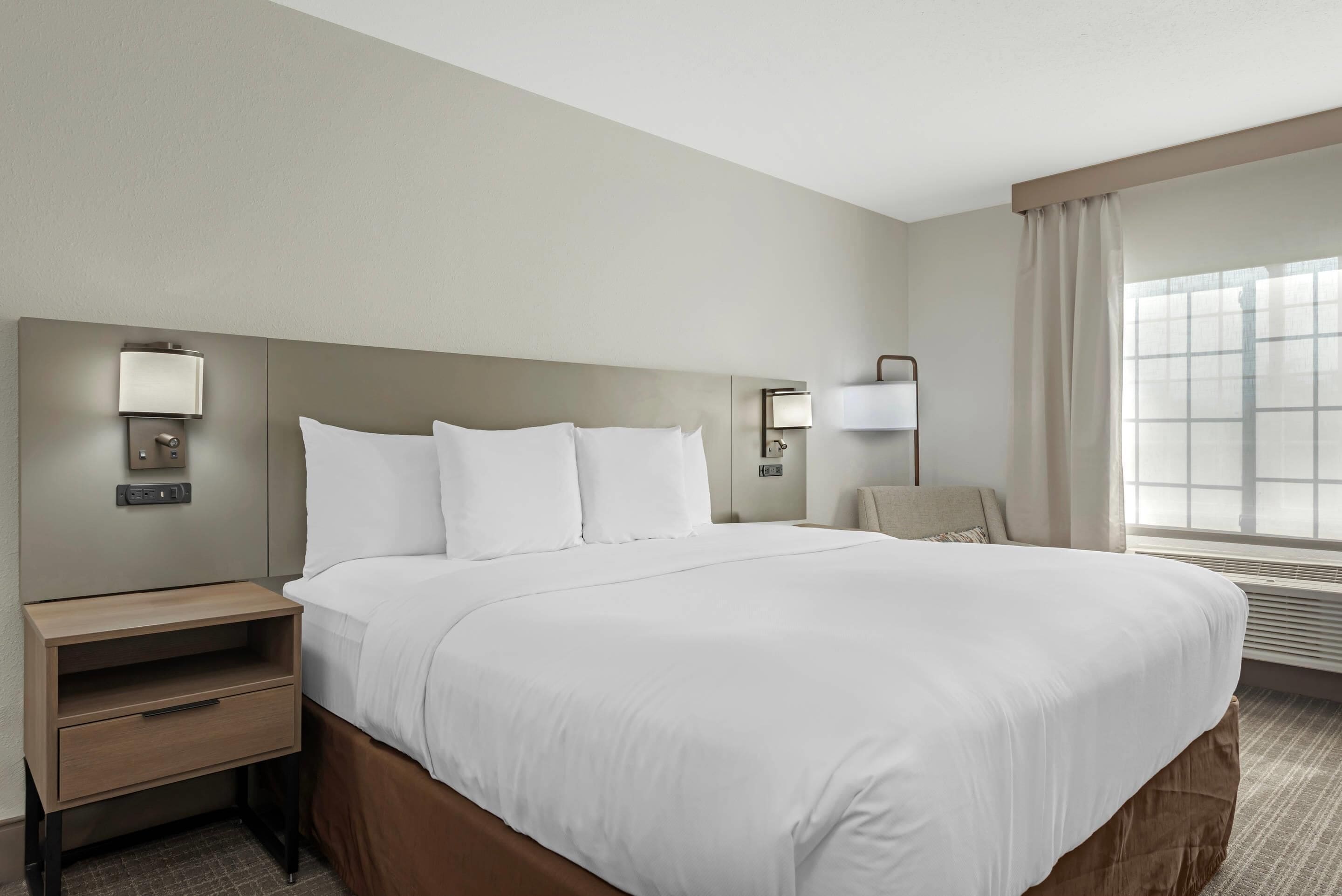 Habitación de hotel con una cama grande, una mesita de noche, una lámpara y una ventana con cortinas.