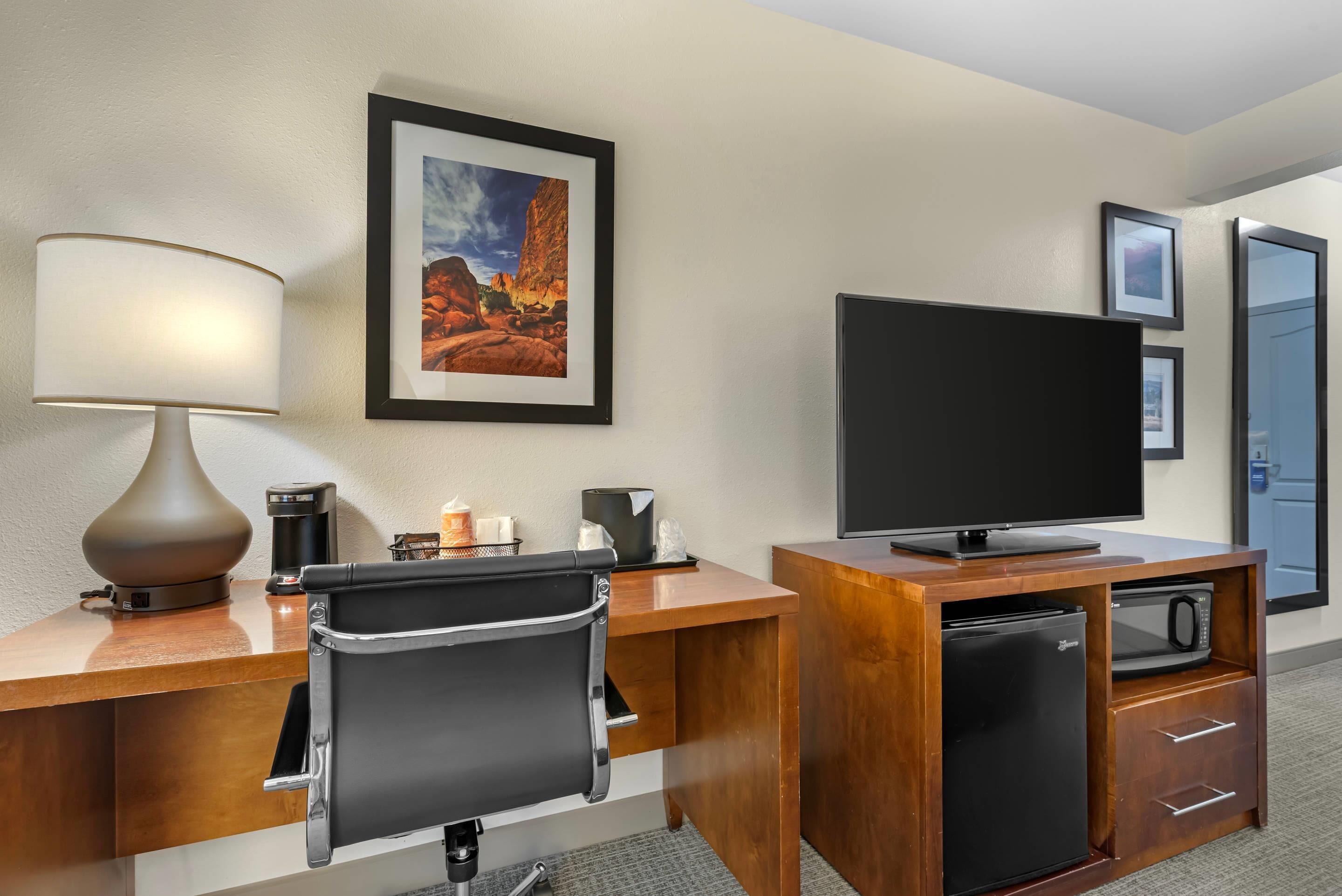 Une chambre d’hôtel avec un bureau en bois, un fauteuil de bureau noir, un téléviseur à écran plat, un mini-réfrigérateur, une machine à café et un tableau représentant un paysage, accroché au mur.