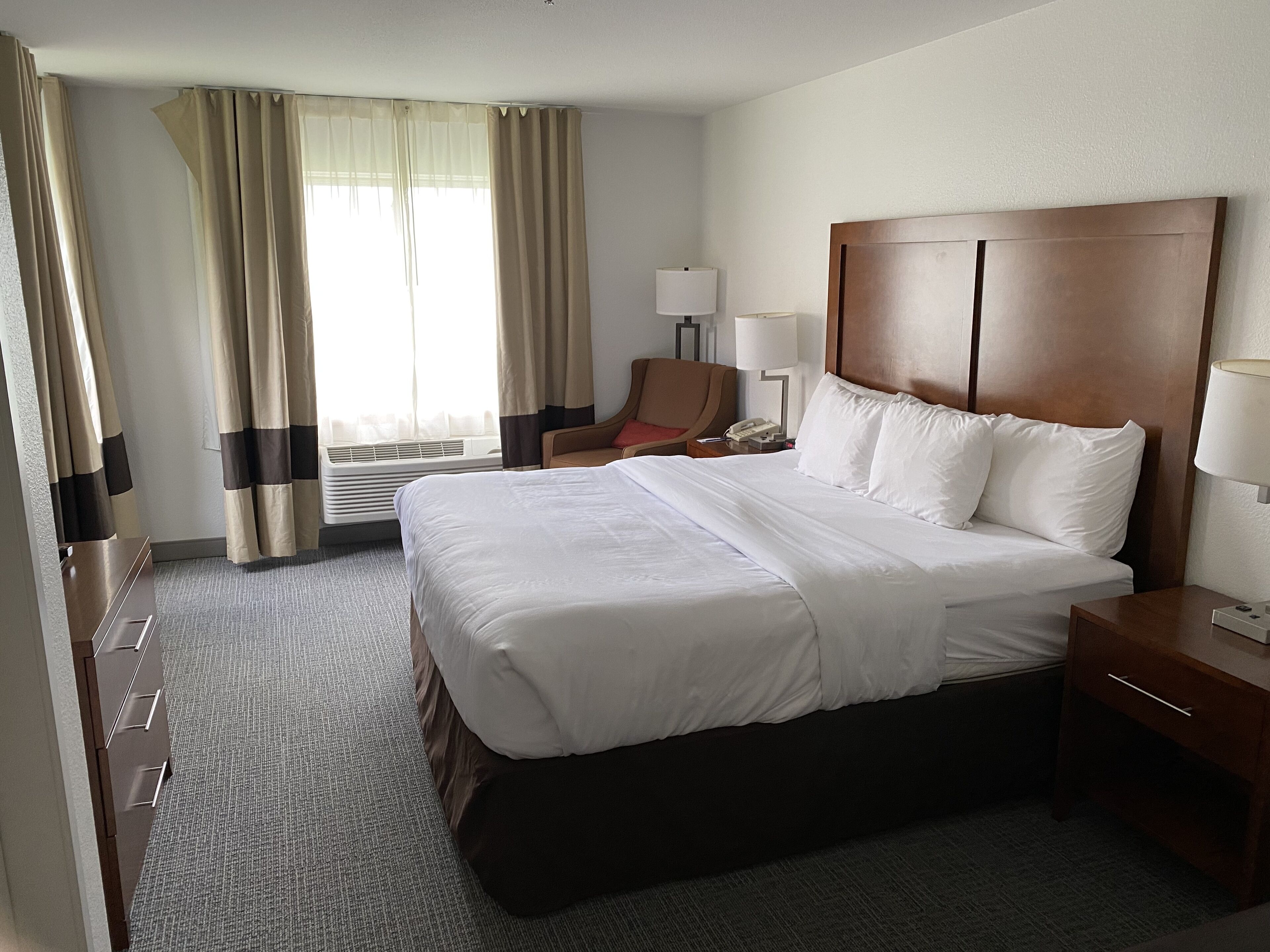 A hotel room with a large bed, a wooden headboard, a desk with a lamp, a chair, and a window with curtains.