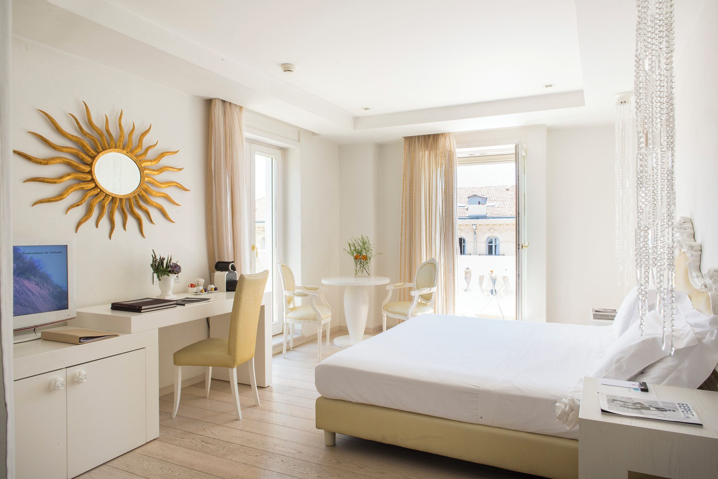 A hotel room with a bed, desk, chairs, a round table, a sun-shaped mirror, and a view of the outside.