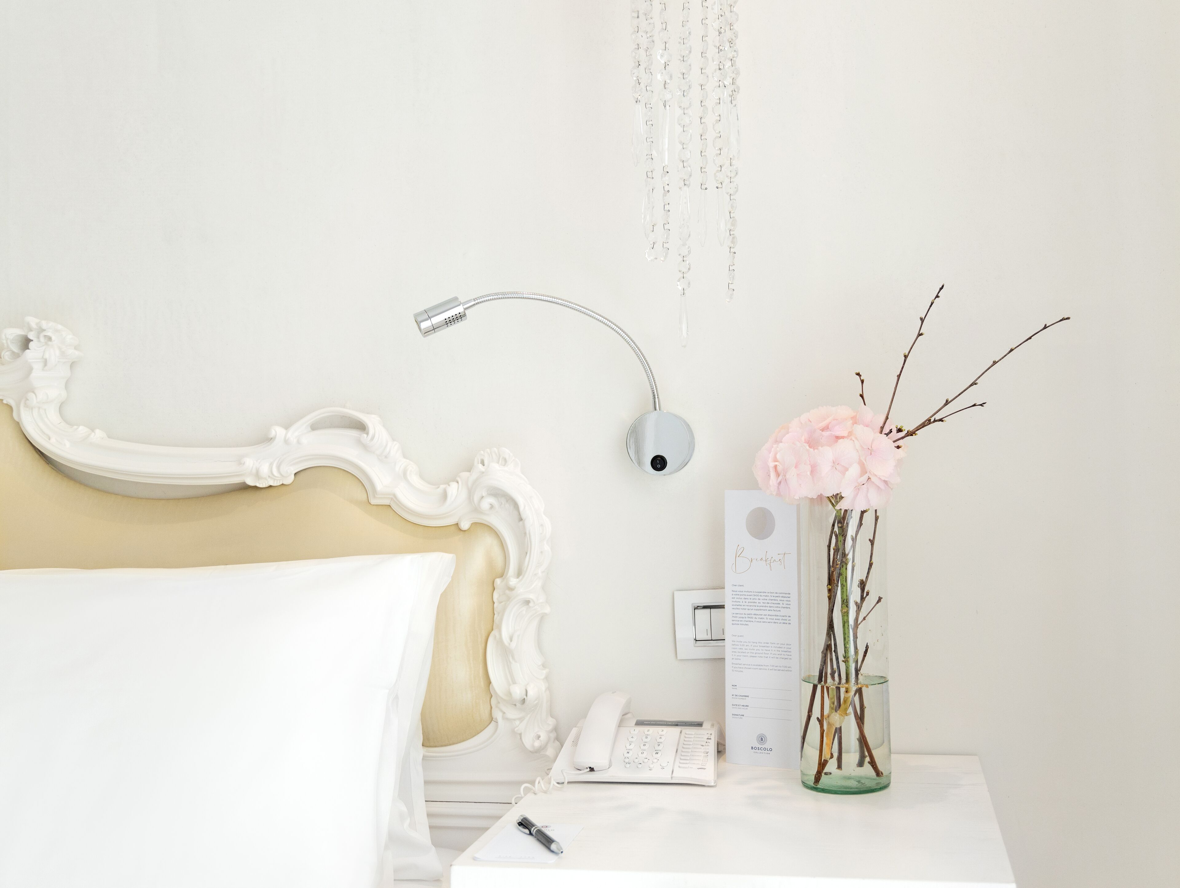 Habitación de hotel impecable con una cama blanca clásica, una mesita de noche con lámpara, un jarrón con ramas y un teléfono.