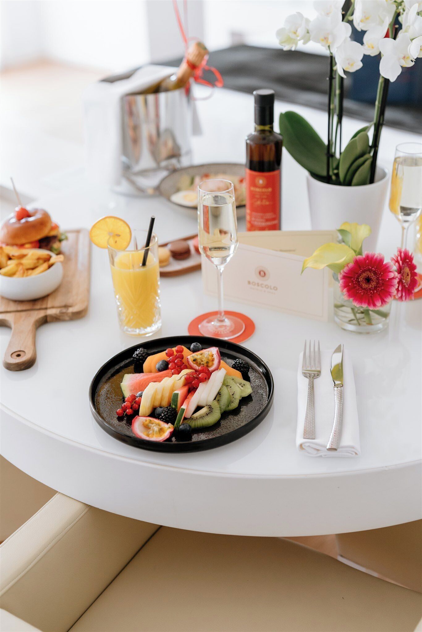 A dining table with a fruit platter, a glass of orange juice, a bottle of olive oil, a wine glass, and a candle.