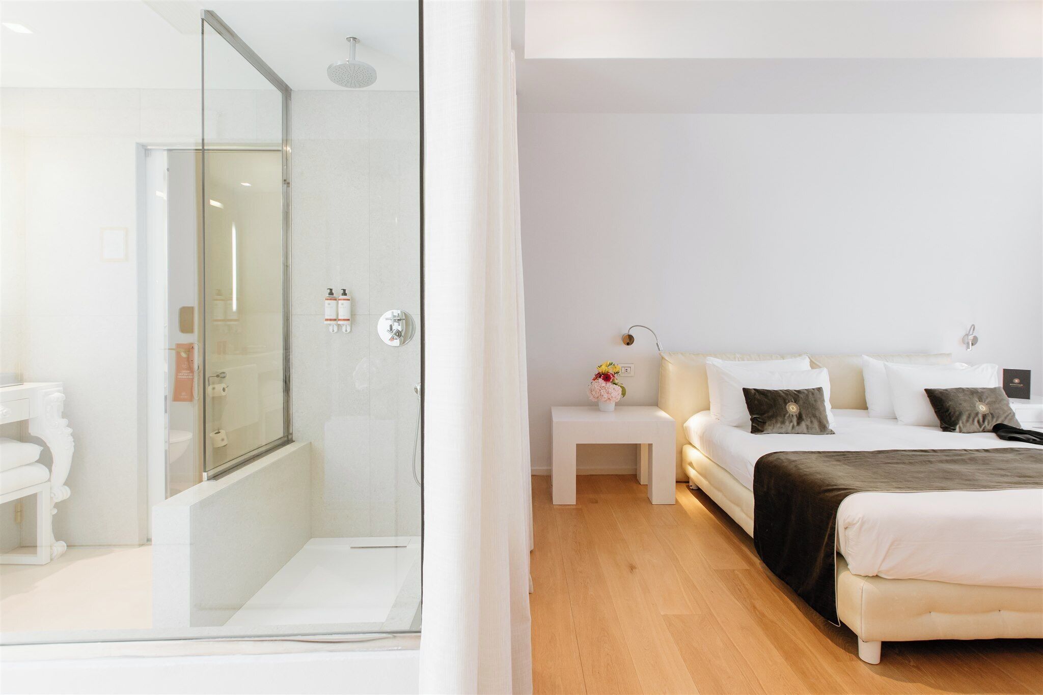 A modern hotel room with a glass-enclosed shower, a bed with white bedding and a dark throw, a small bedside table with a vase, and a wooden floor.