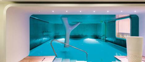 Piscina interior moderna com uma estrutura em forma de árvore, três cadeiras de descanso com toalhas e um vaso branco grande.