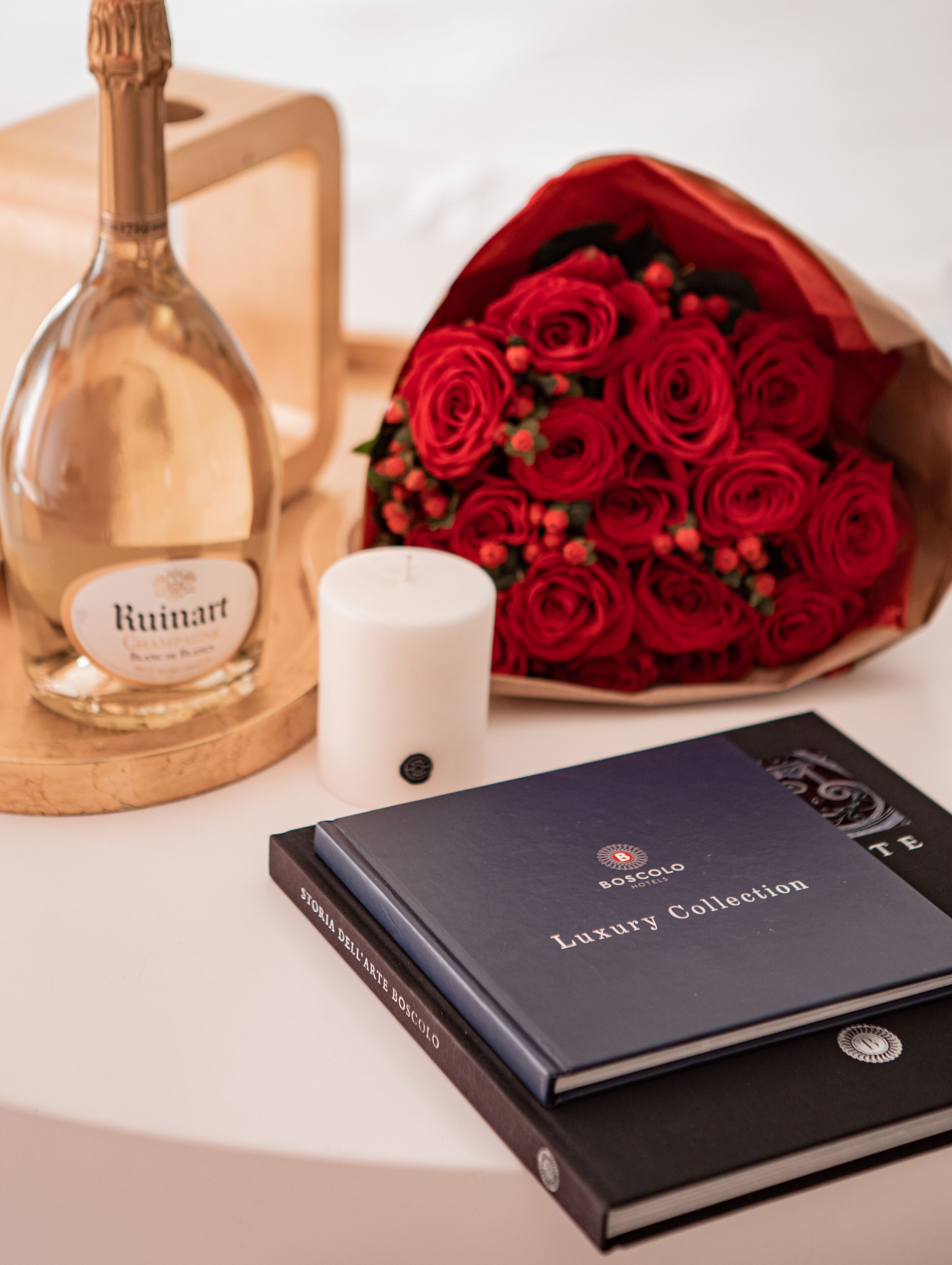 A bottle of Ruinart champagne, a white candle, a bouquet of red roses, and a book titled 'BOSCOLO Luxury Collection'.
