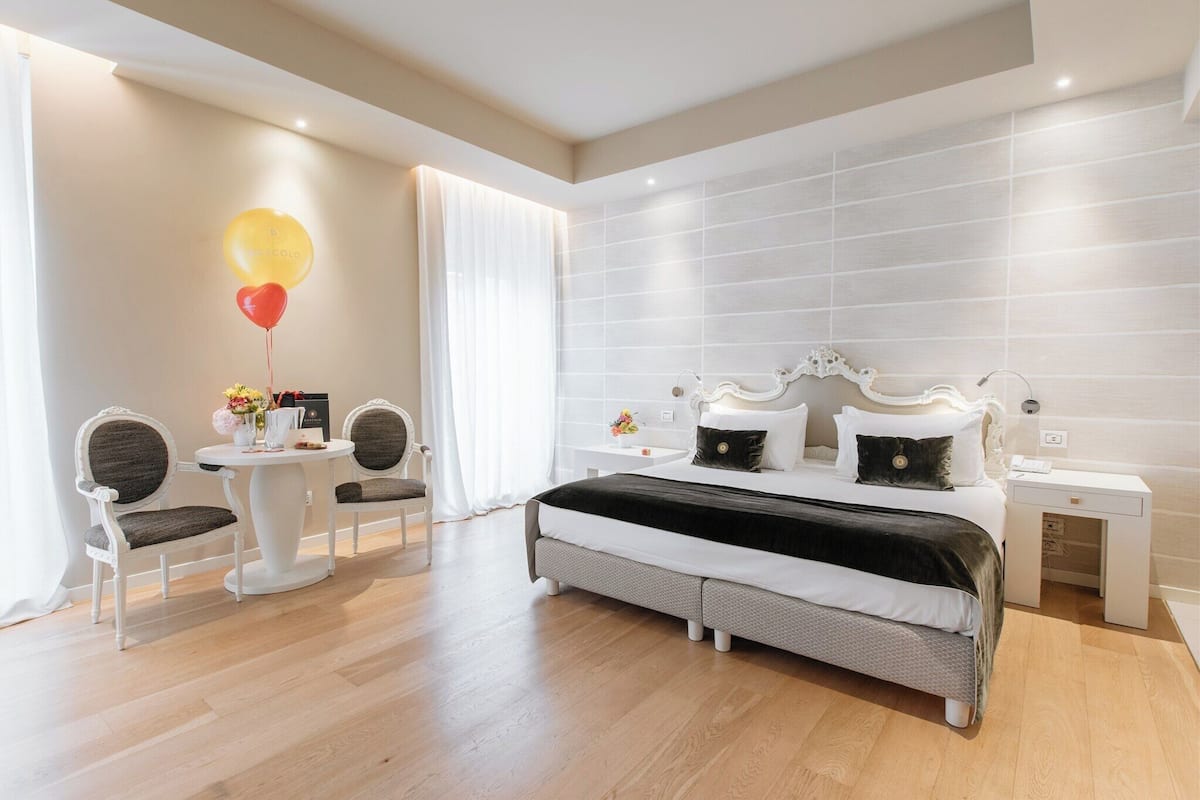 A modern hotel room with a large bed, a bedside table, a chair, a small table with flowers, and a window with curtains.