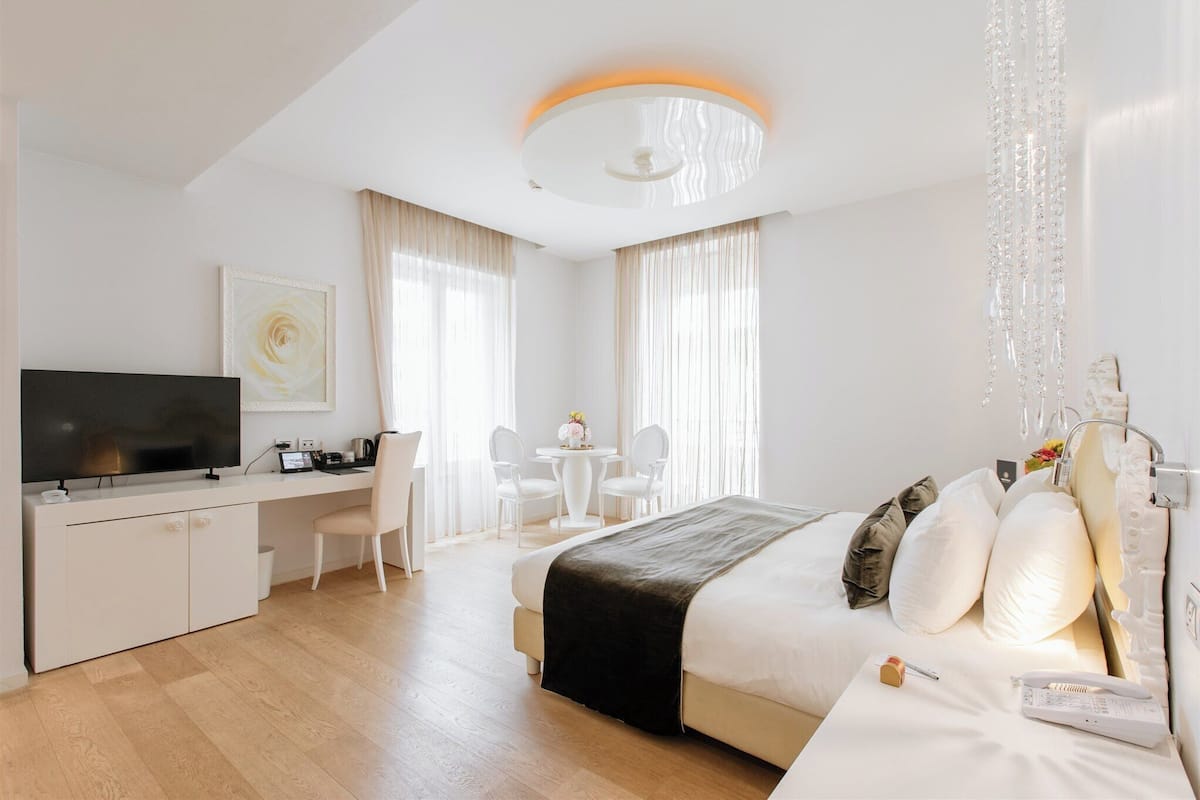 A modern hotel room with a bed, bedside table, and a desk with a chair. There is a television on the desk, a window with curtains, and a chandelier.