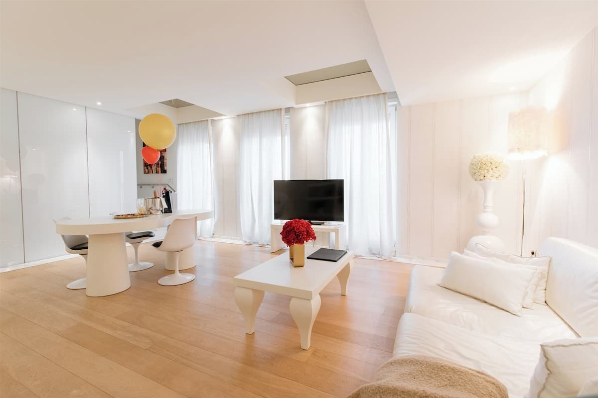 A modern living room with a white sofa, a dining table with chairs, a flat-screen TV, and a white lamp.