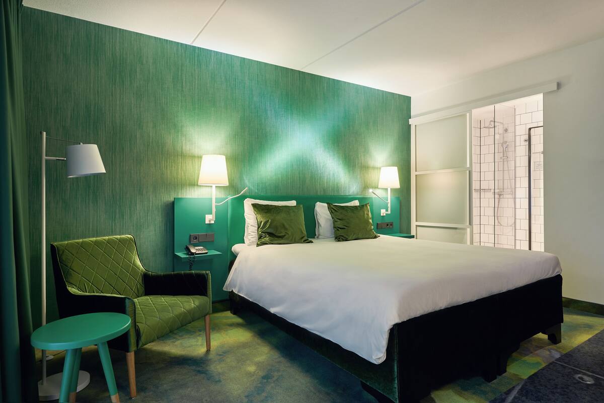 A hotel room with a large bed, two bedside lamps, a green upholstered chair, a small round table, and a view of a tiled shower area.