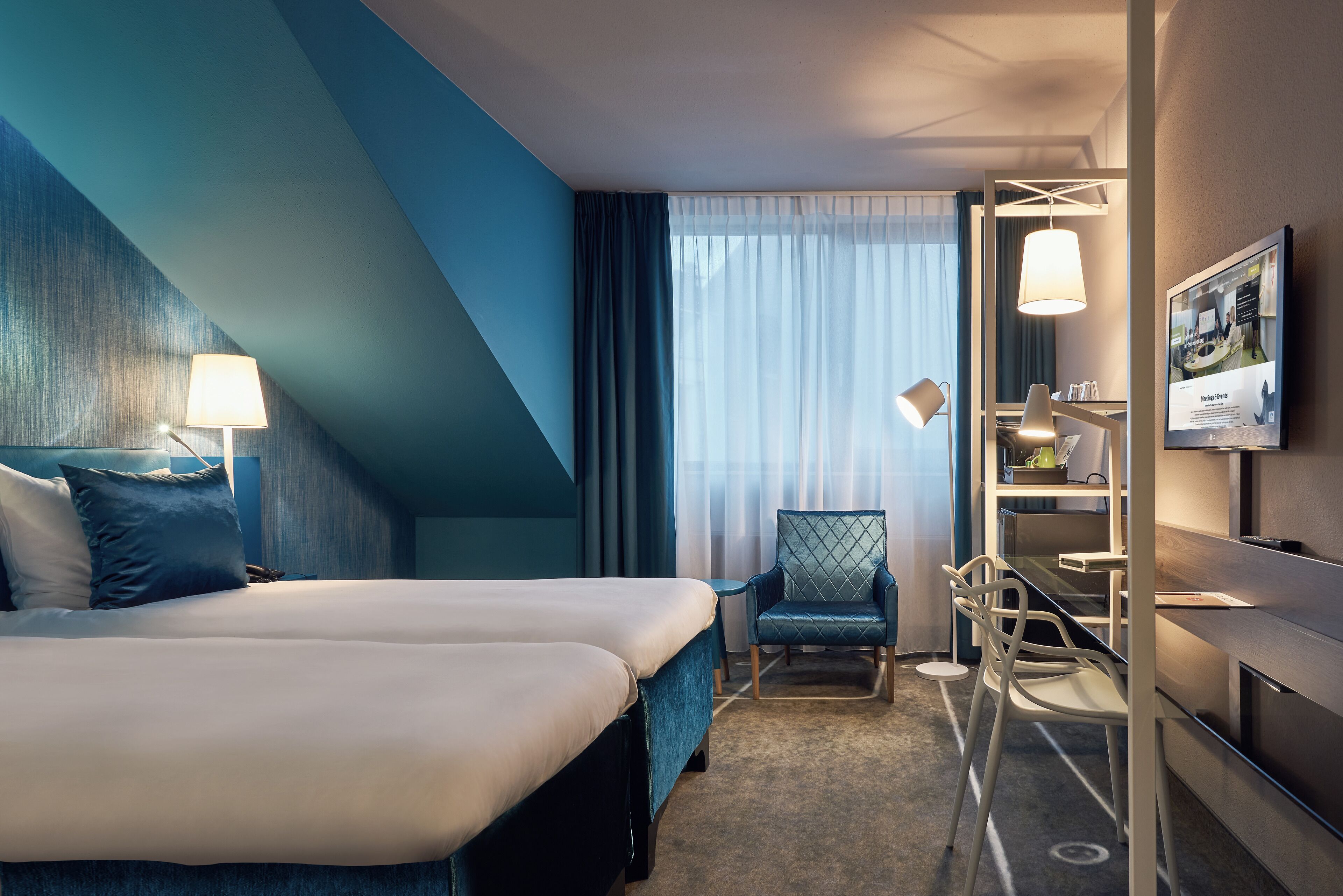 Une chambre d’hôtel avec un lit, un bureau, une chaise, une télévision et une fenêtre avec des rideaux.