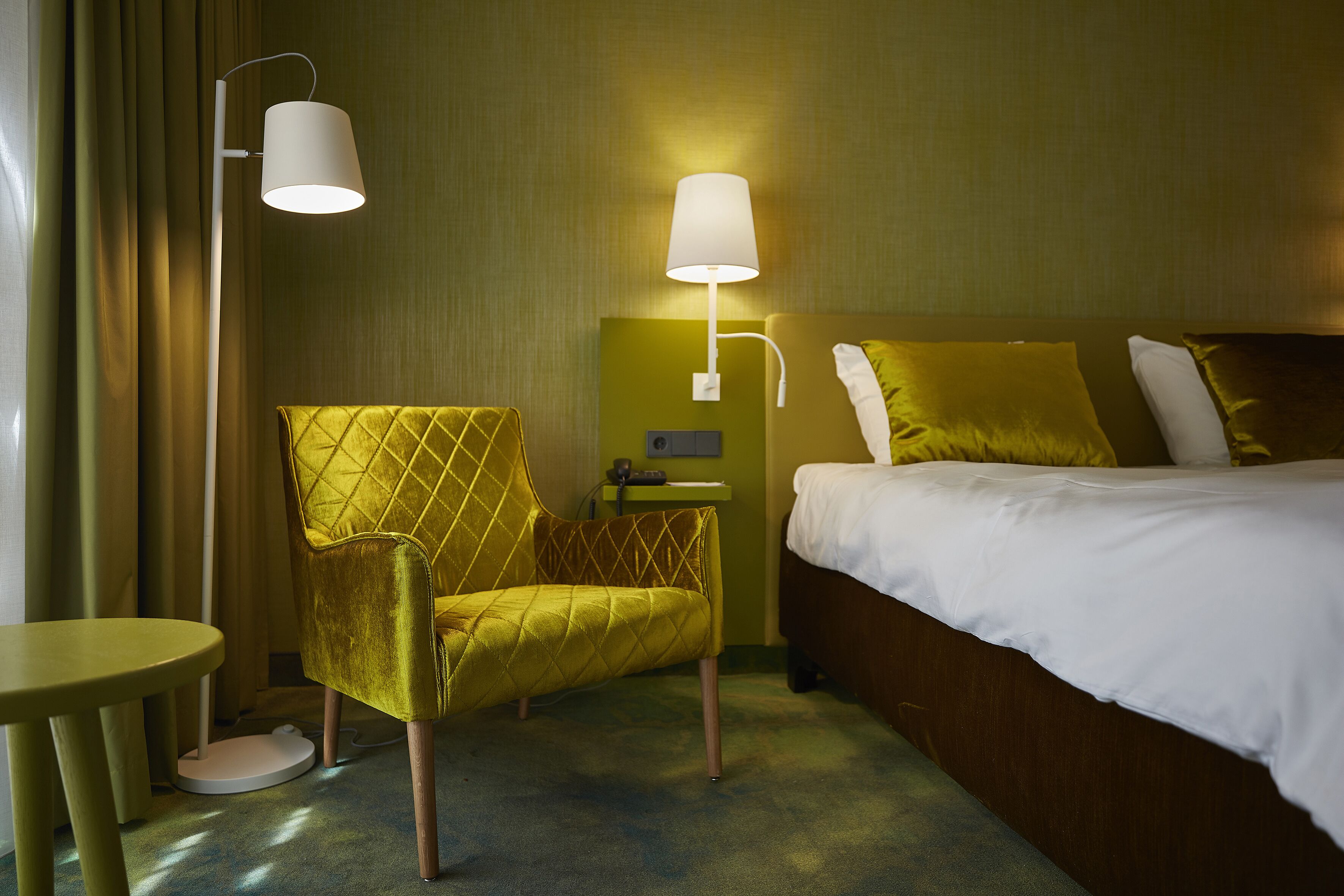Une chambre d’hôtel avec un lit, un fauteuil jaune, une table de chevet et deux lampes fixées au mur.