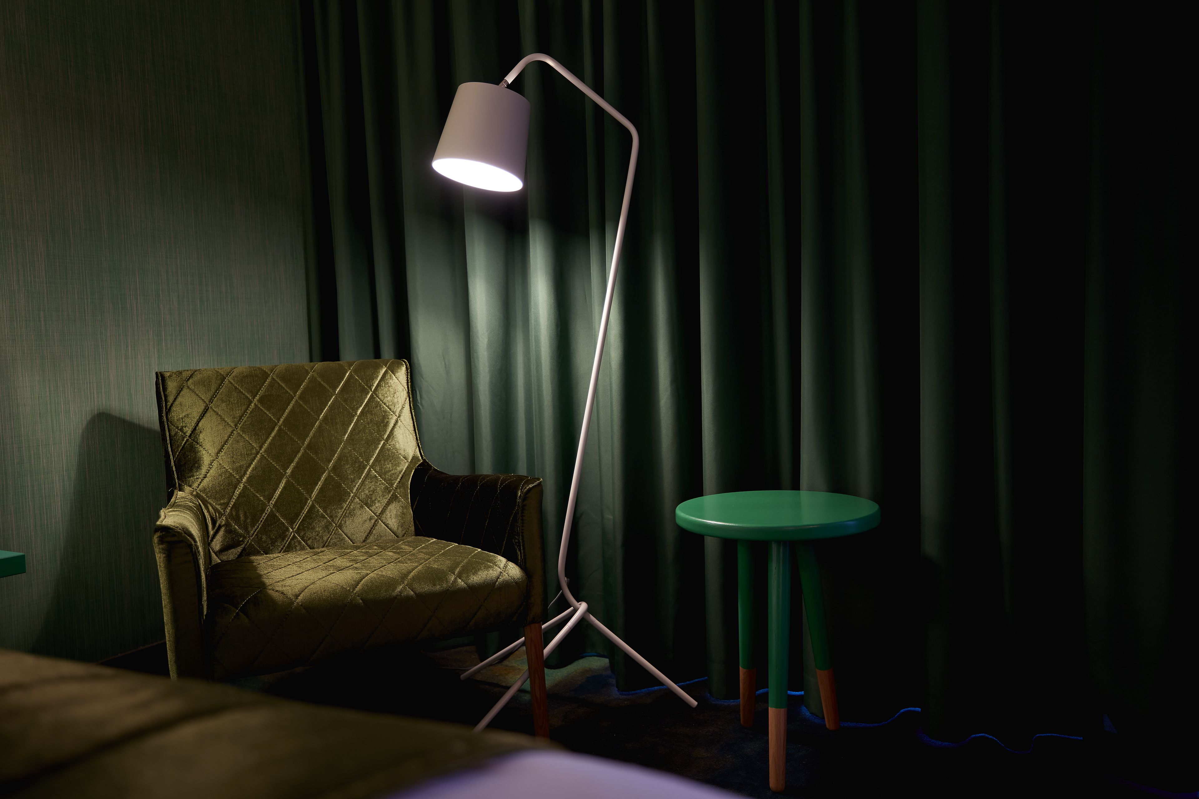 Een kamer met een groene bekleden fauteuil, een vloerlamp en een kleine ronde tafel.