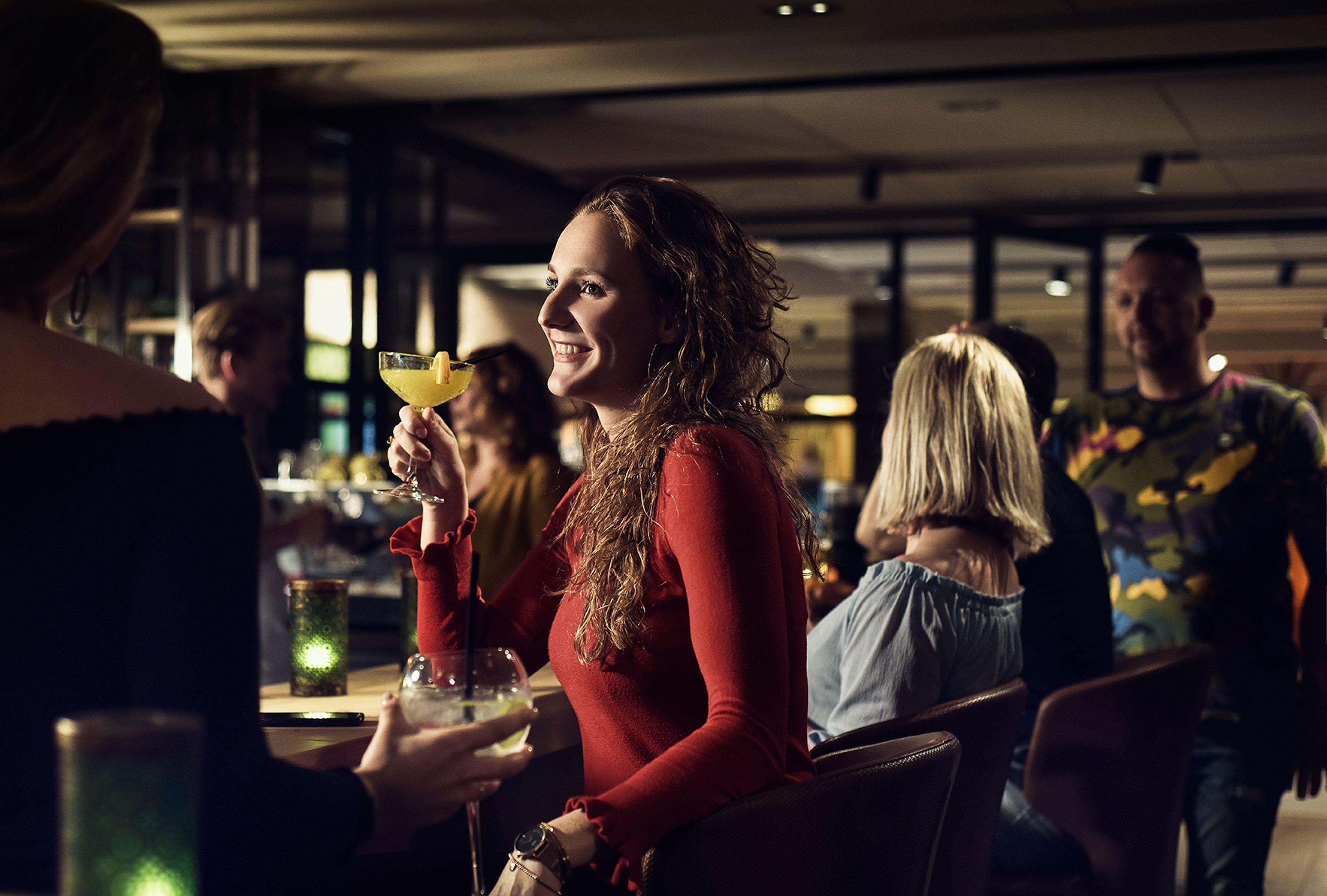 A person holding a drink at a bar with other people in the background.