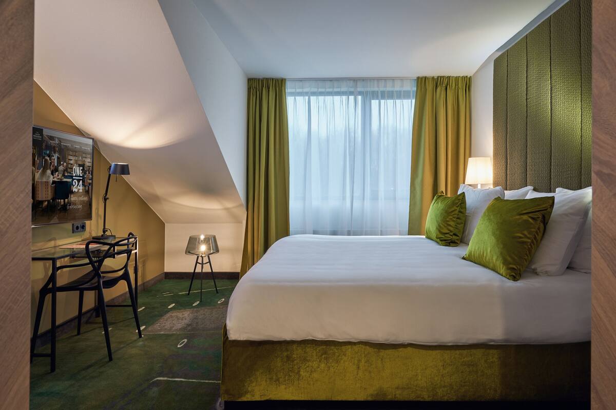 A hotel room with a bed, a desk with chairs, a lamp, and a window with curtains.