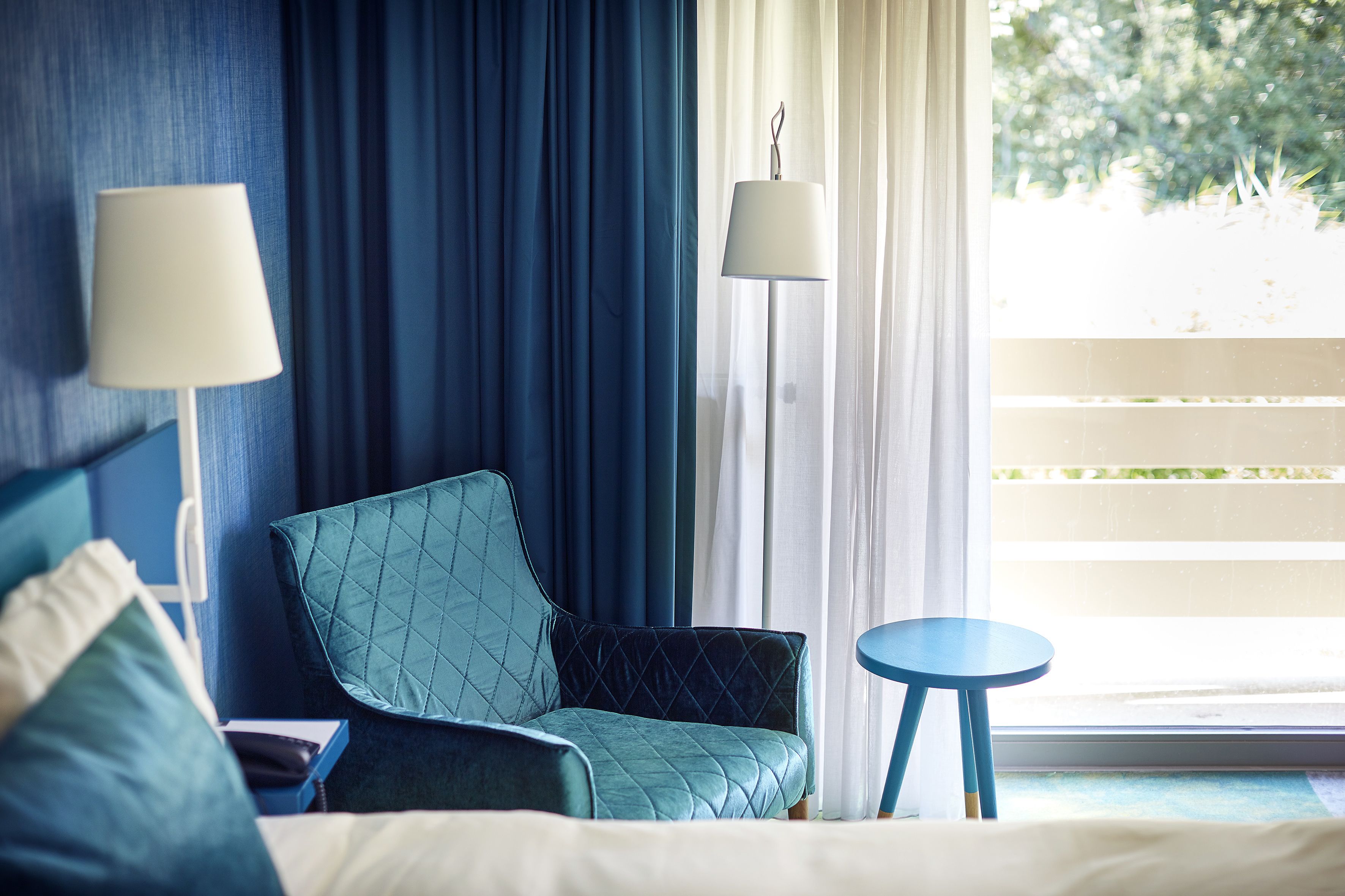 Une chambre d’hôtel avec un lit, un fauteuil bleu, une petite table ronde et une lampe sur pied.