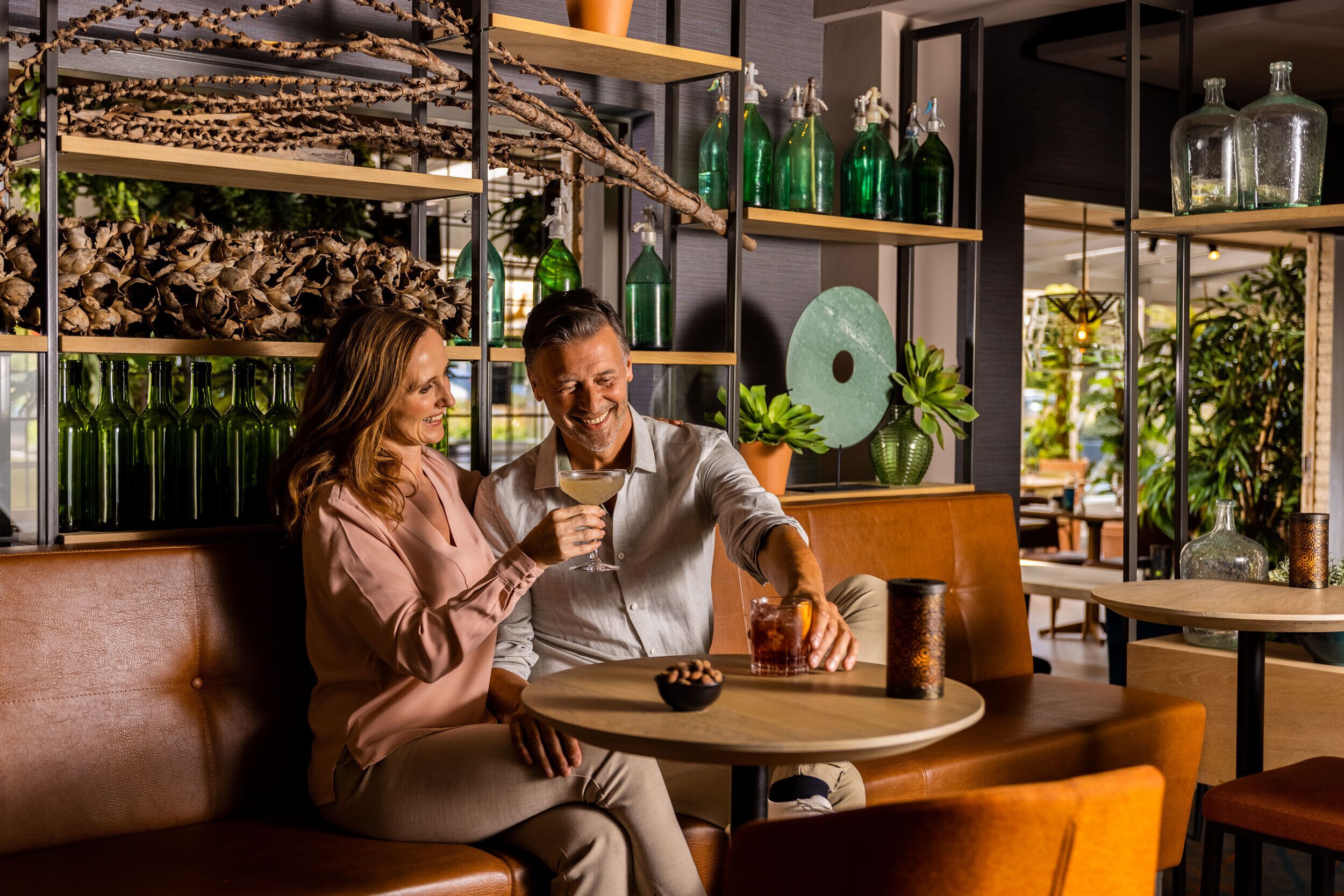 A couple seated on a leather couch in a modern interior with shelves of green bottles and plants.