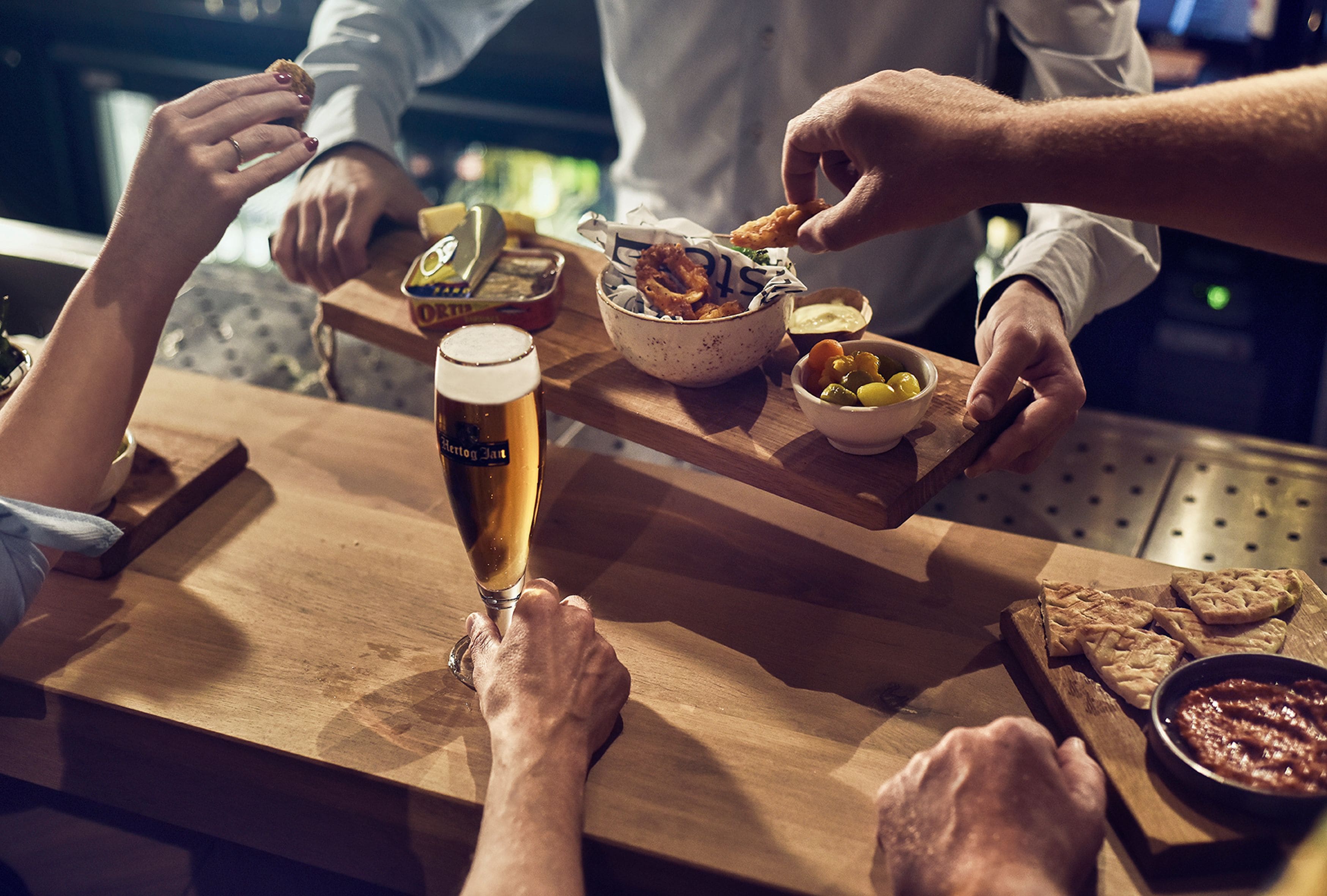 A wooden bar counter with a glass of beer, bowls of snacks, and a person serving food.