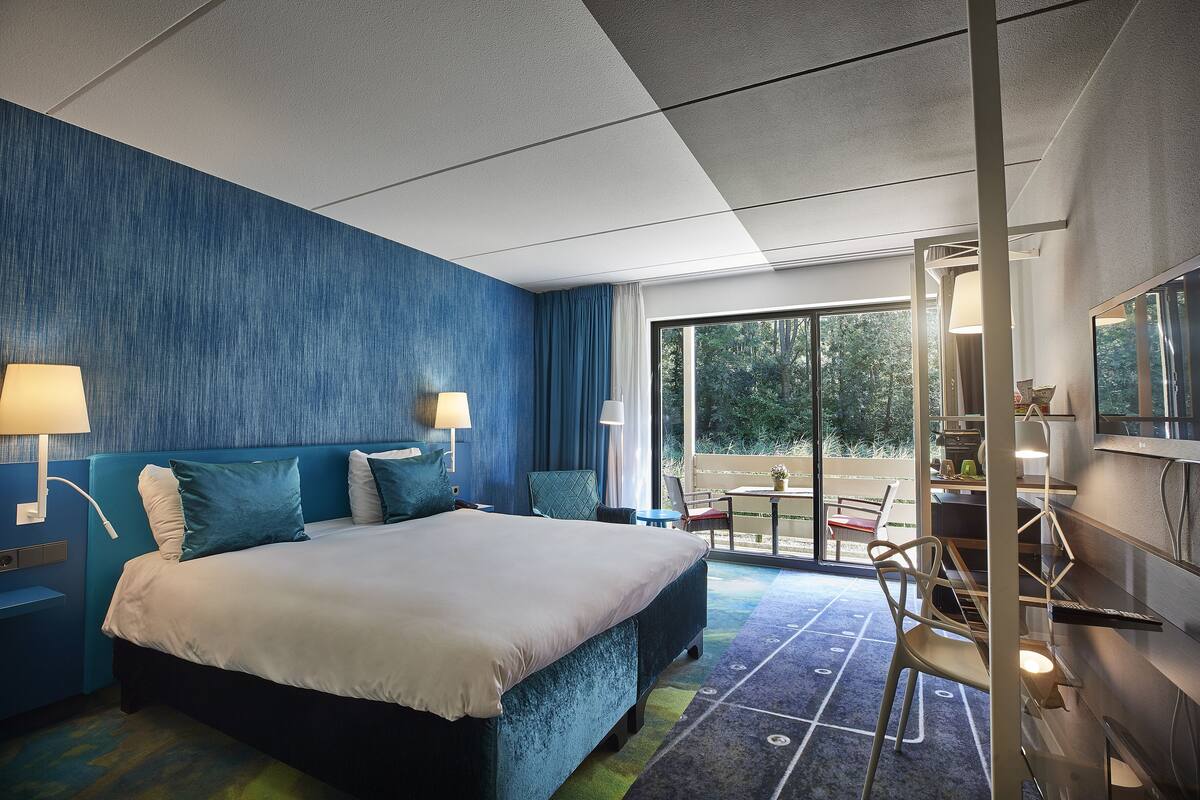 A modern hotel room with a large bed, two bedside lamps, a desk with a chair, and a view of the outdoors through a large window.