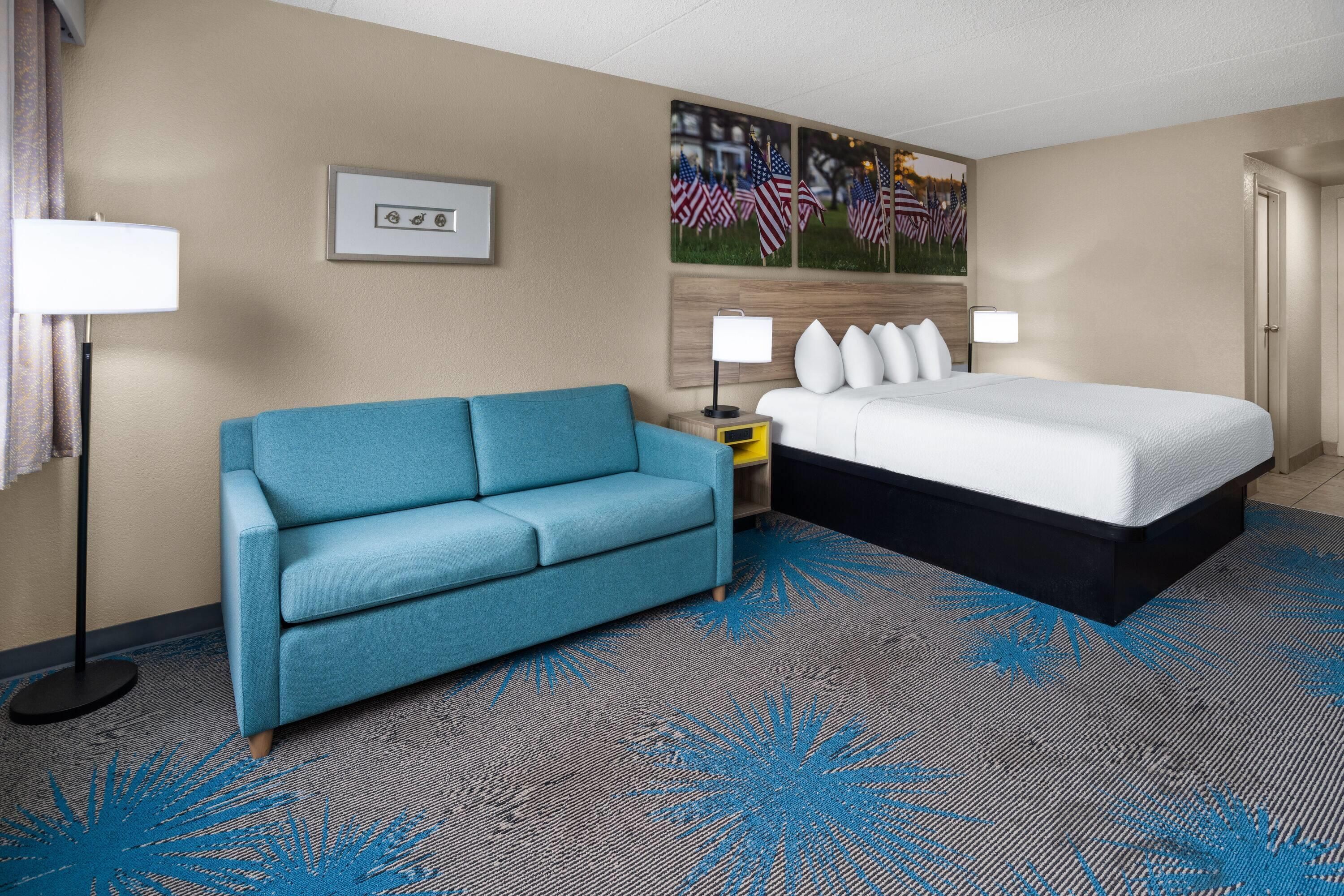 A hotel room with a bed, a blue sofa, a nightstand, a lamp, and a painting on the wall.