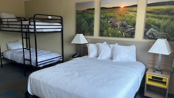 Une chambre d’hôtel avec un lit superposé, un lit simple, deux lampes de chevet, une table de chevet et un tableau représentant un coucher de soleil sur un champ.