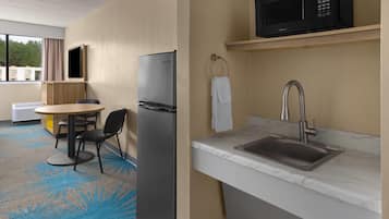 Habitación de hotel compacta con kitchenette, zona de comedor con mesa y sillas, ventana grande y televisor de pantalla plana montado en la pared.