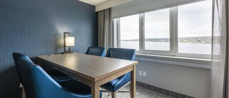 A hotel room with a wooden table and blue chairs, a window with a view of a cityscape, and a lamp on the table.
