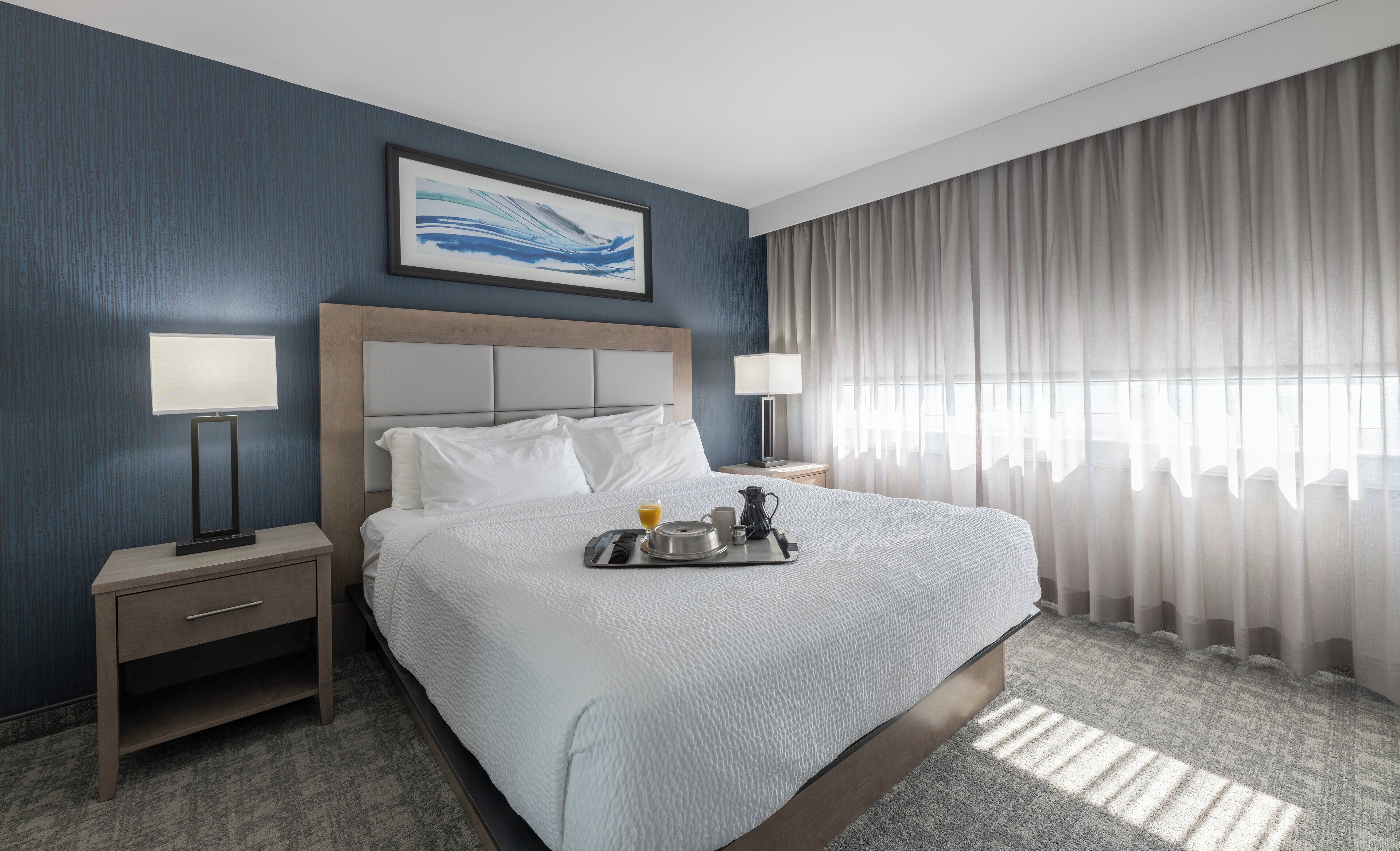 Habitación de hotel con una cama grande, dos mesitas de noche, una lámpara de noche, un cuadro enmarcado sobre la cama y una ventana con cortinas.