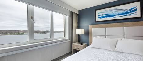 A hotel room with a large window, a bed with white linens, a nightstand with a lamp, and a framed abstract painting on the wall.