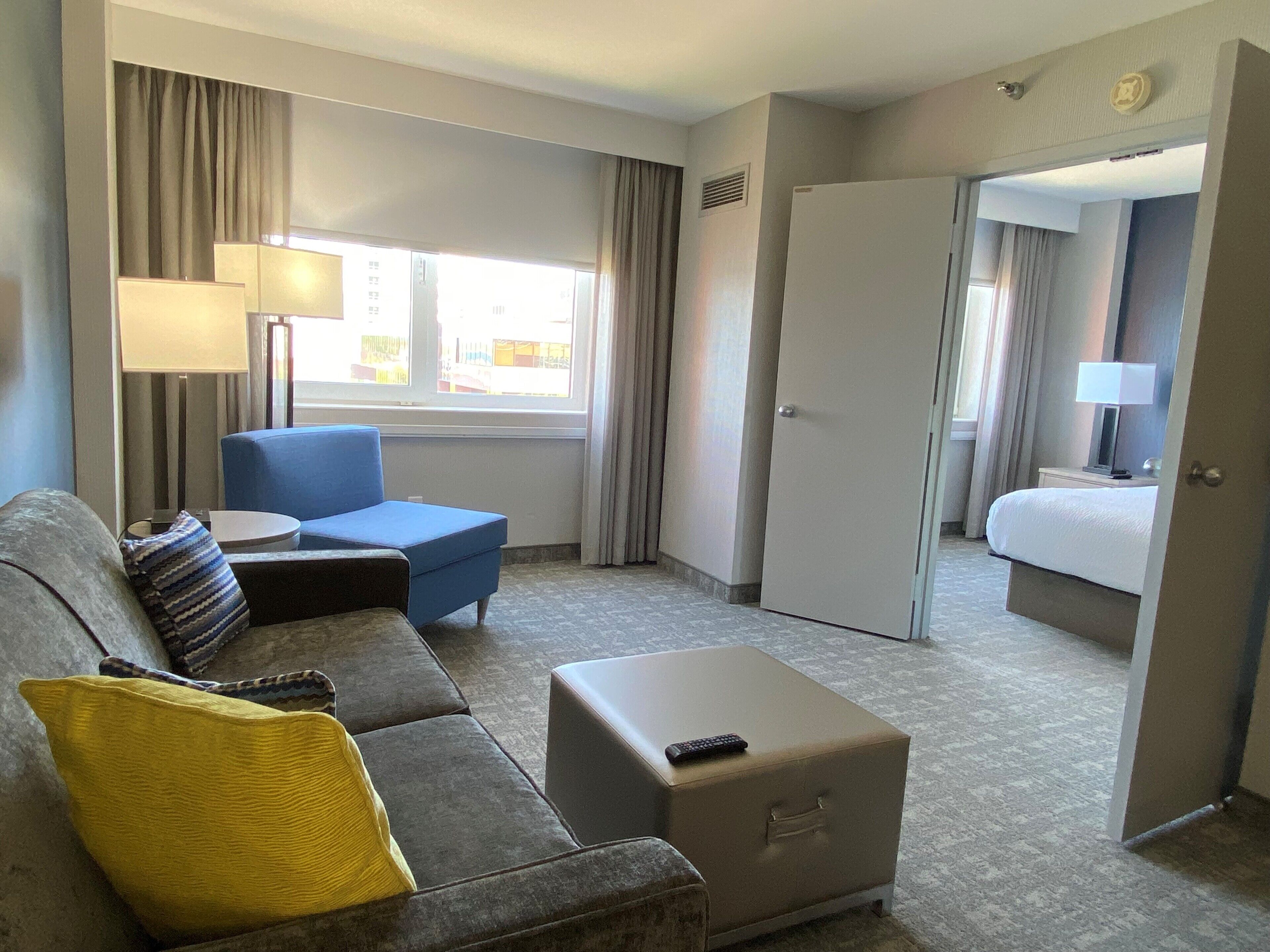 Ein Hotelzimmer mit grauem Sofa, blauem Sessel, kleinem Hocker, Nachttisch mit Lampe, Bett und einer Tür zu einem weiteren Raum.