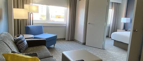 Ein Hotelzimmer mit grauem Sofa, blauem Sessel, kleinem Hocker, Nachttisch mit Lampe, Bett und einer Tür zu einem weiteren Raum.