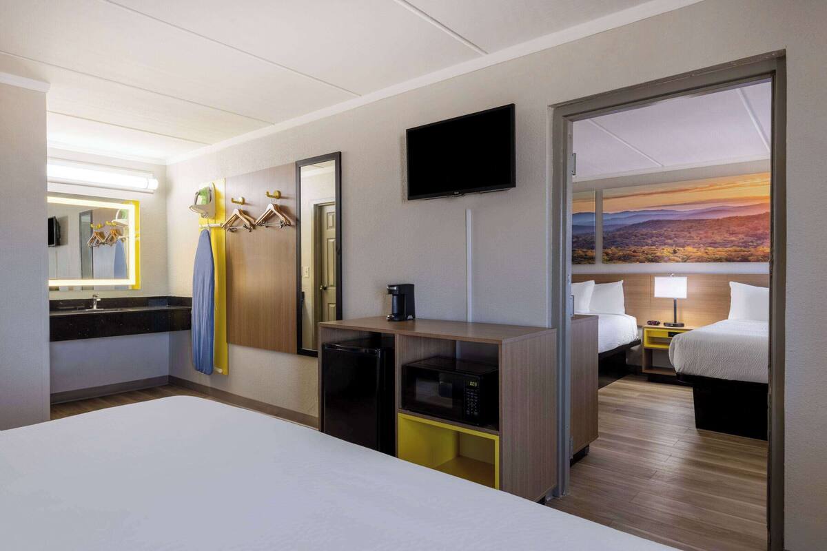 Une chambre d’hôtel équipée d’un lit, d’un bureau avec un ordinateur, d’un four à micro-ondes, d’une machine à café, d’une télévision, d ’un miroir et d’un tableau représentant un coucher de soleil.