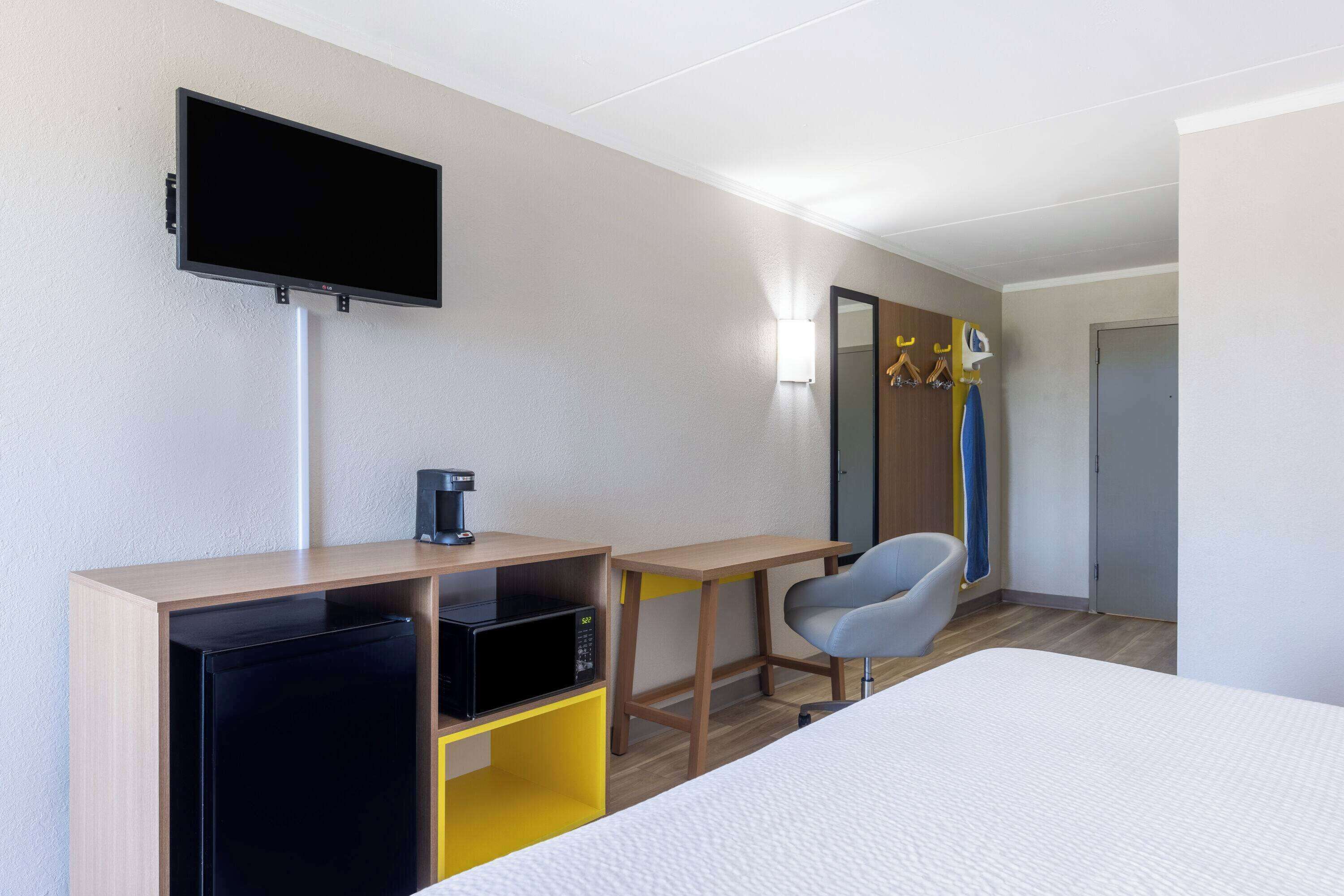 A hotel room with a bed, a desk with a computer, a chair, a TV mounted on the wall, and a door.