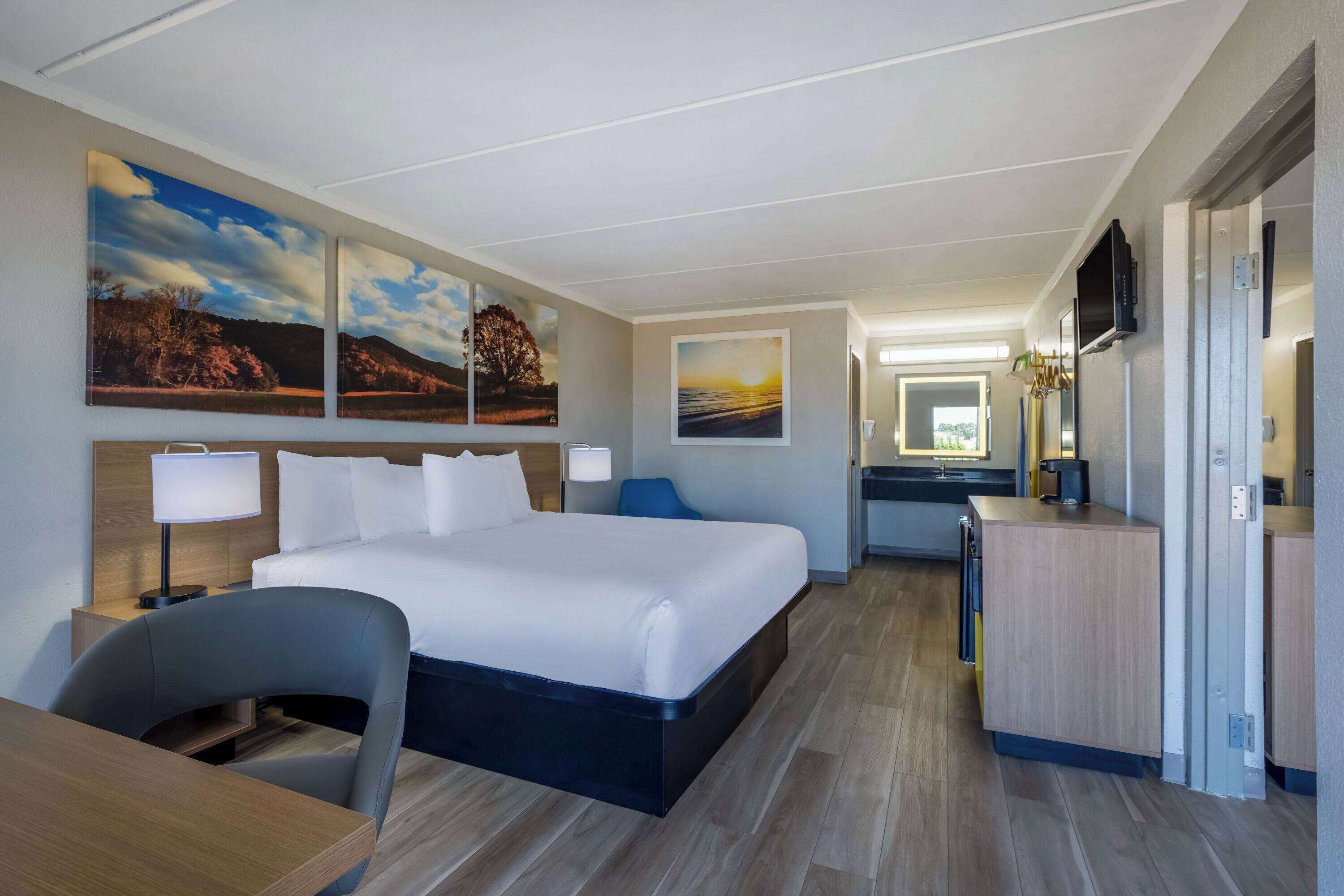A hotel room with a large bed, a desk, a chair, and a TV. There are paintings on the wall and a desk lamp.
