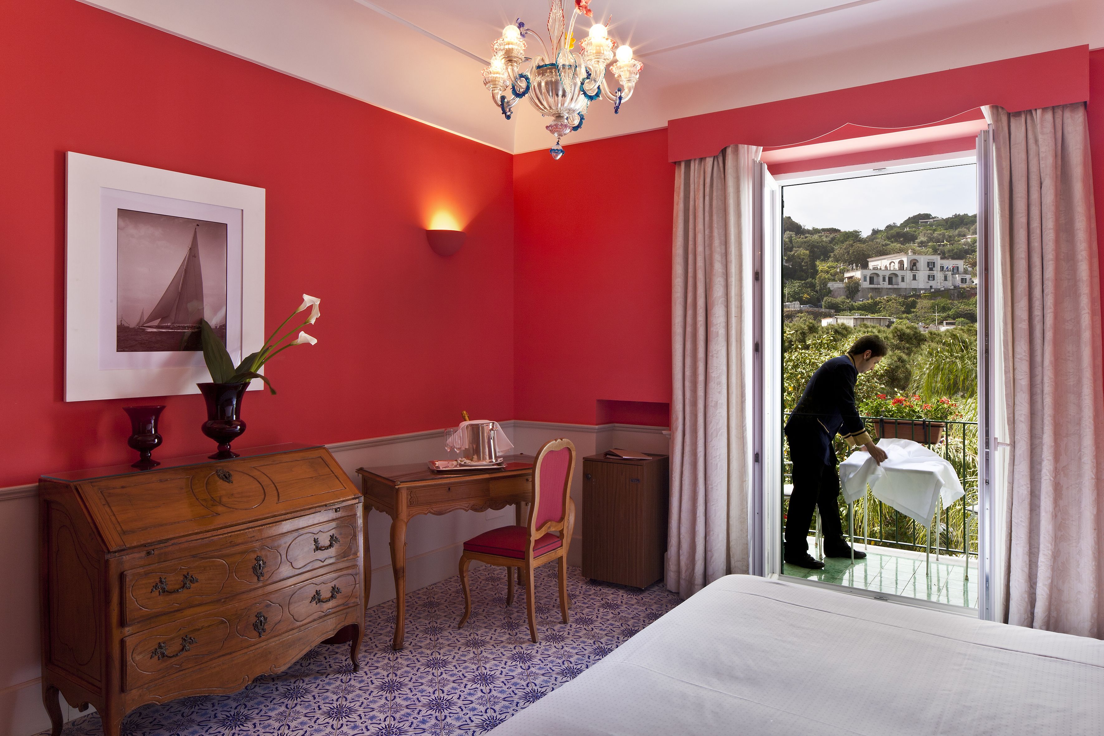 A hotel room with a red wall, a wooden desk, a chair, a dresser, and a bed. There is a window with a view of a building and greenery outside.