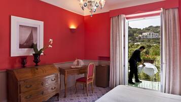 A hotel room with a red wall, a wooden desk, a chair, a dresser, and a bed. There is a window with a view of a building and greenery outside.