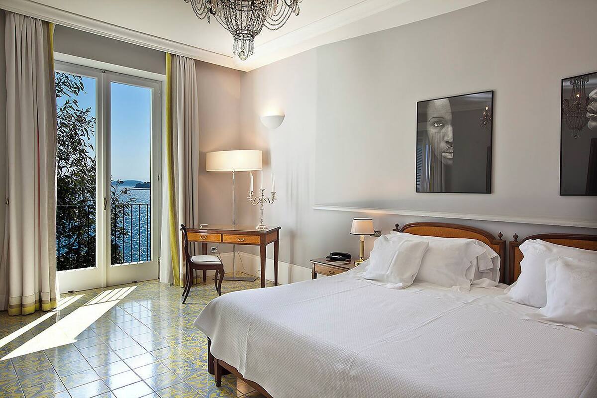 Une chambre d’hôtel avec un grand lit, un bureau, une chaise, une table de chevet, une lampe et une vue sur la mer depuis un balcon.