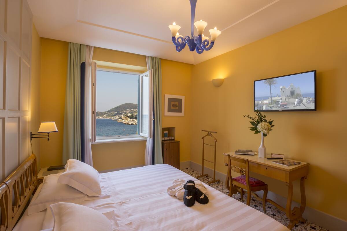 Une chambre d’hôtel avec un lit, un bureau, une chaise, une fenêtre donnant sur la mer et un lustre.