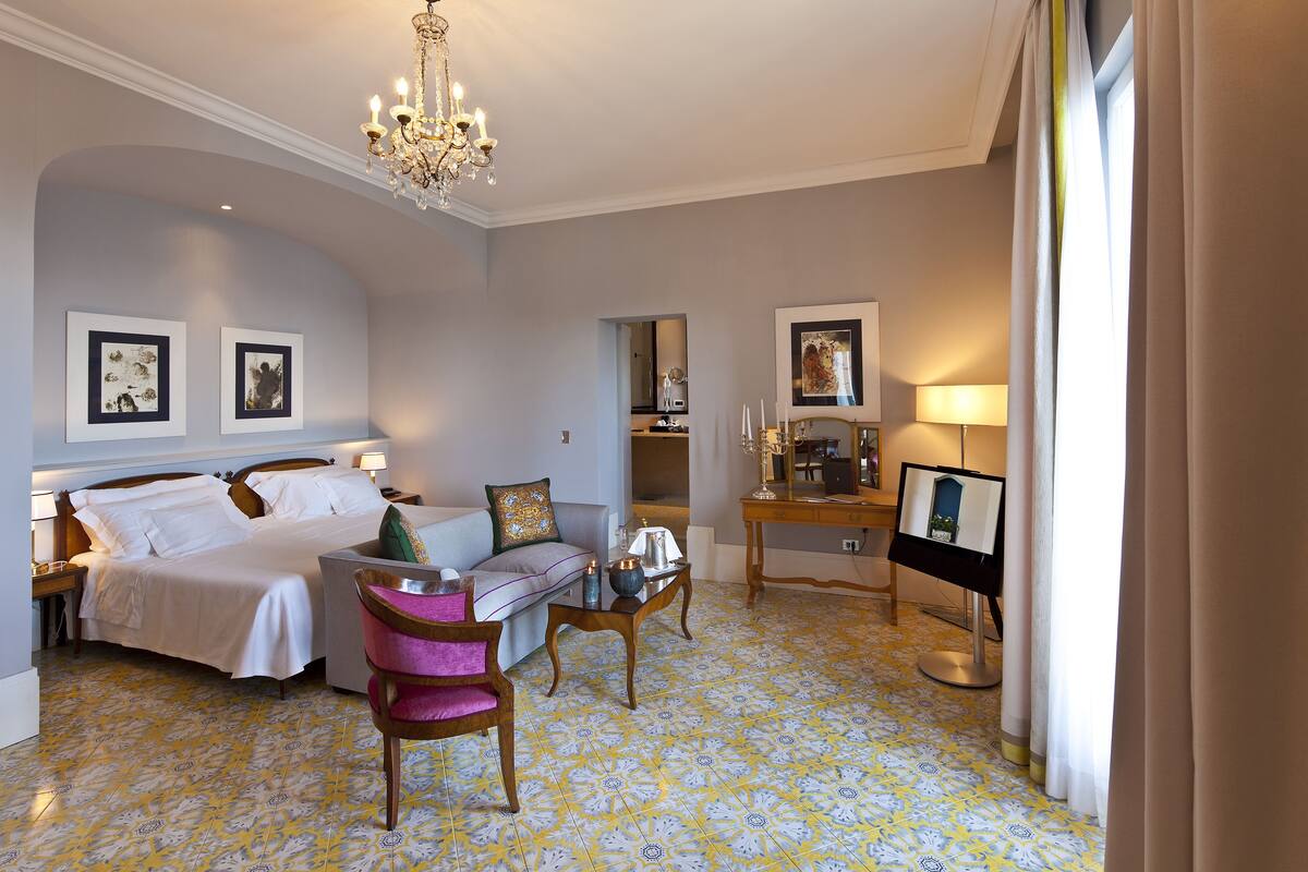 Une chambre d’hôtel comprenant un lit, un canapé, une chaise, une petite table, un lustre et des œuvres d’art accrochées aux murs.