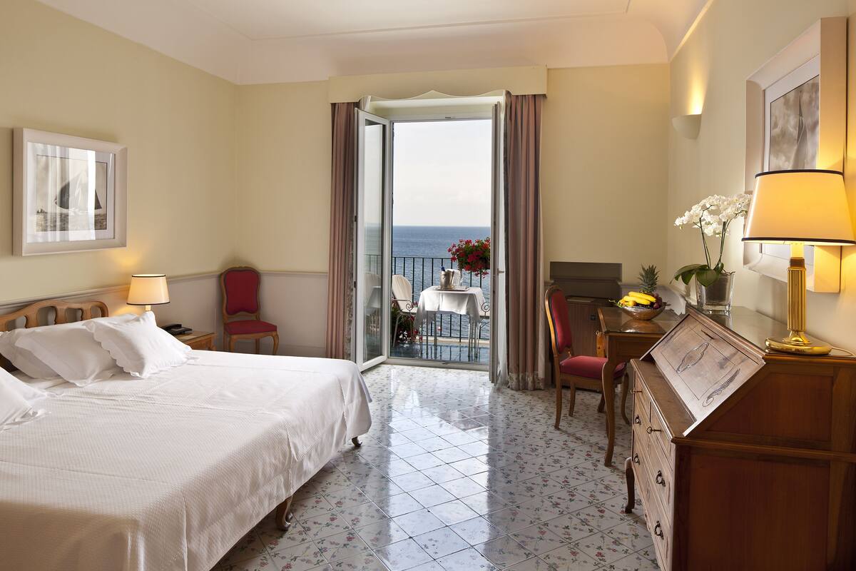 Une chambre d’hôtel avec un lit, un bureau, une chaise, une table ornée de fleurs et une vue sur la mer, visible par une porte ouverte.