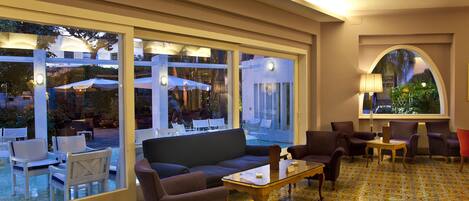 Un área de estar interior con un sofá, sillas y una mesa de centro. Amplias ventanas ofrecen vistas del patio exterior.