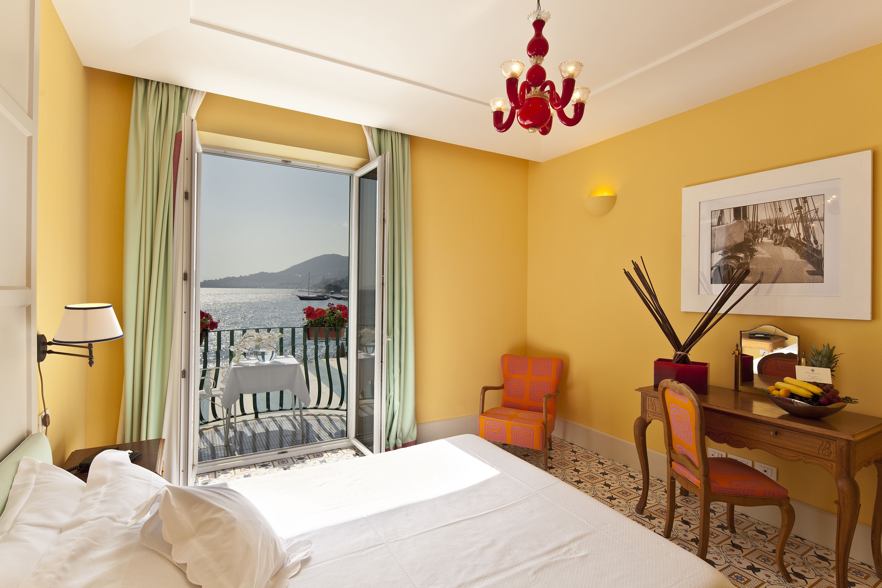 Una habitación de hotel con cama, escritorio y silla, balcón con mesa y sillas, y vista al mar.
