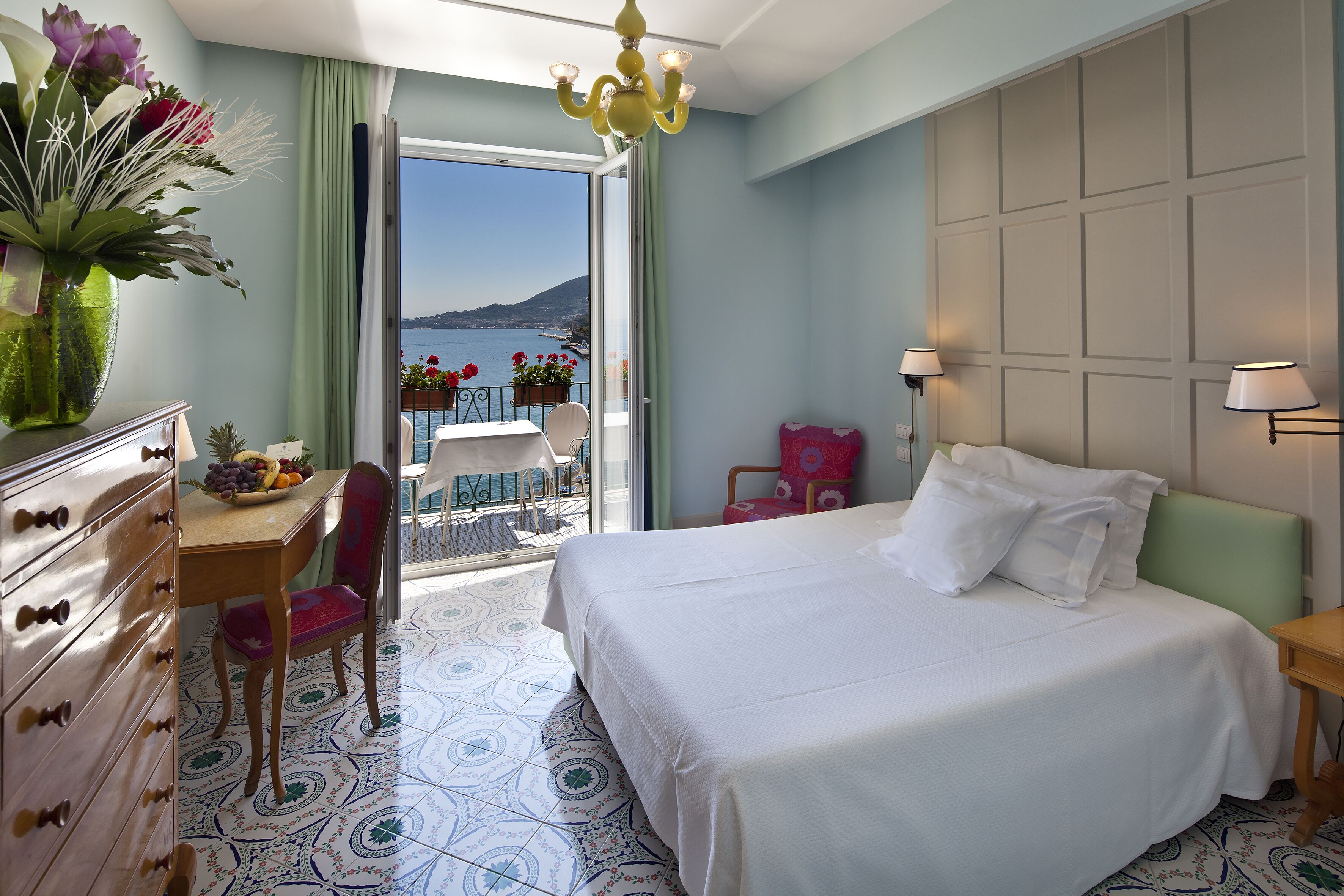 A hotel room with a bed, a desk with a chair, a dresser, a balcony with a table and chairs, and a view of the sea.